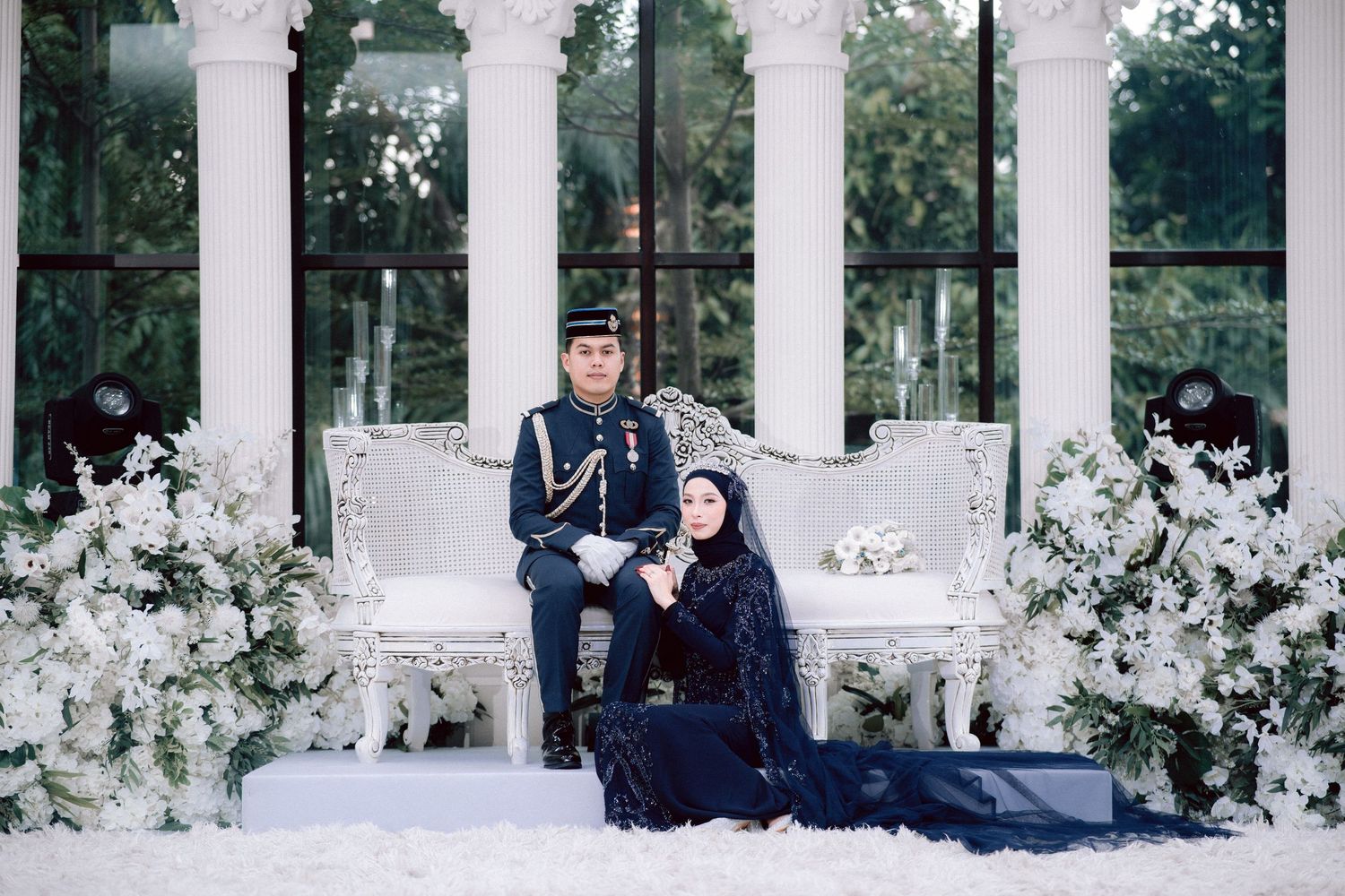 A couple poses in a romantic garden setting with white columns and blooming flowers along a colonnade walkway.