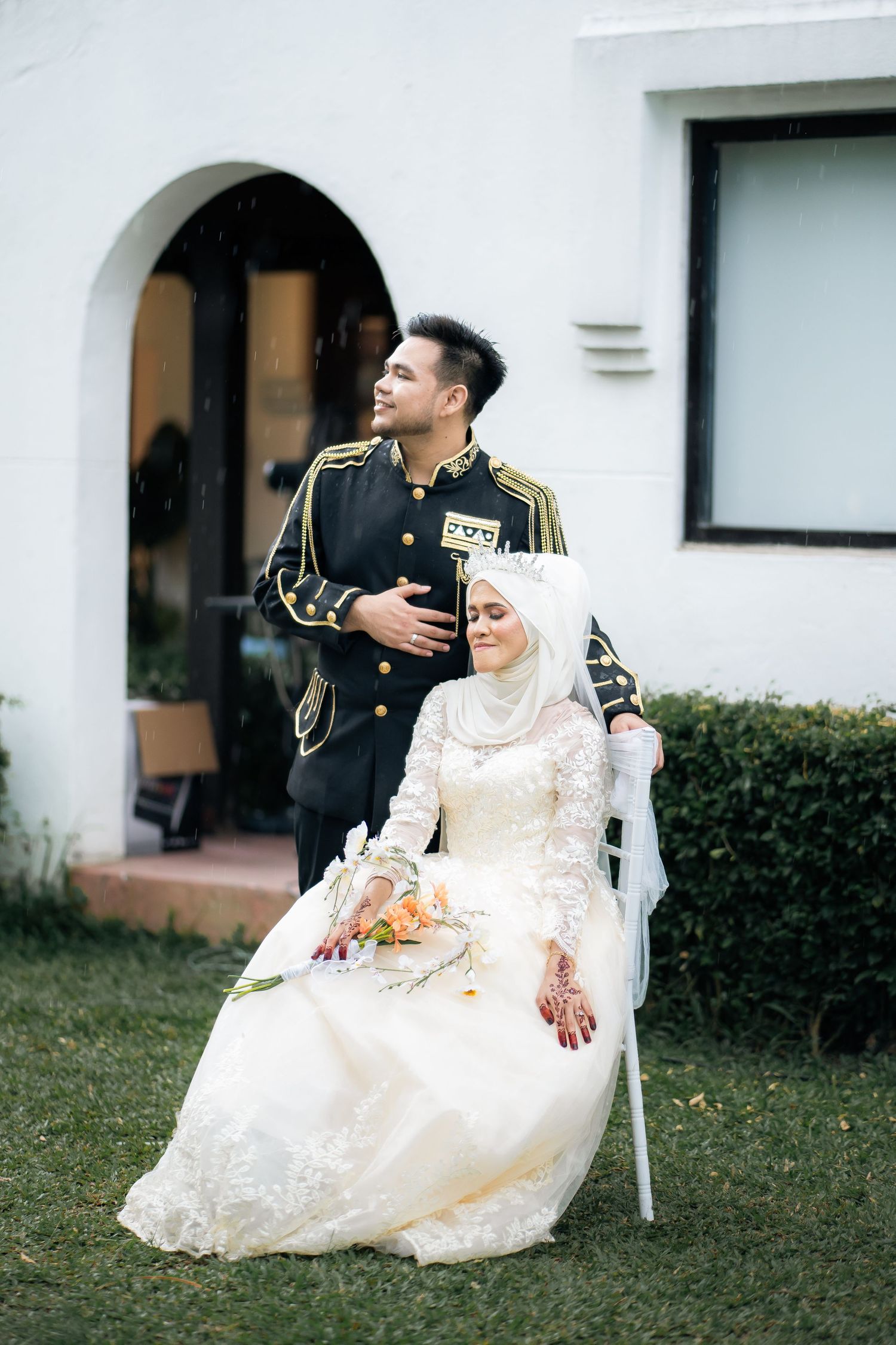 A bride in a white lace dress and hijab sits outdoors while her partner in military uniform stands beside her.