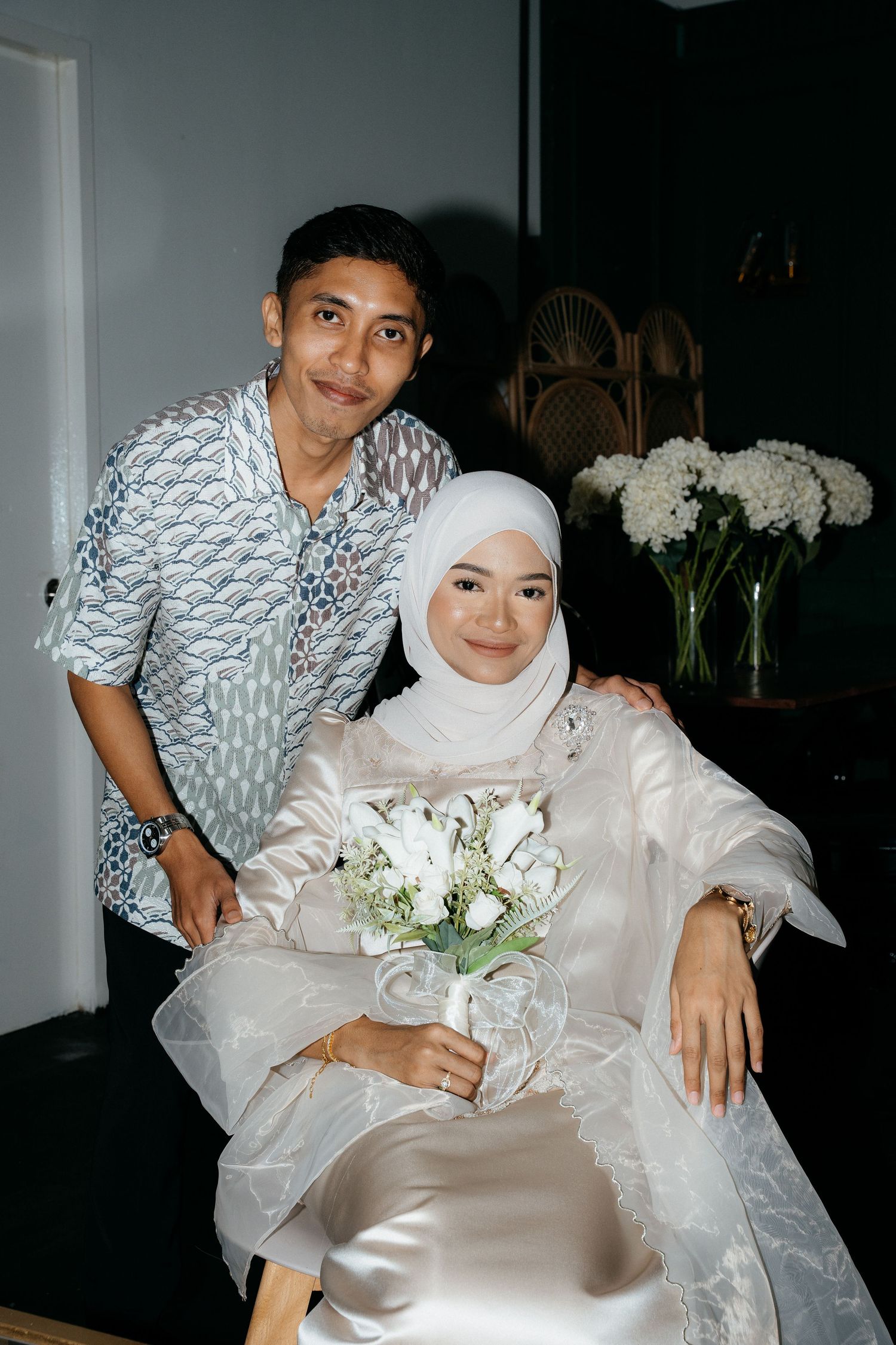Two people in traditional dress pose together with white floral arrangements in a home setting.
