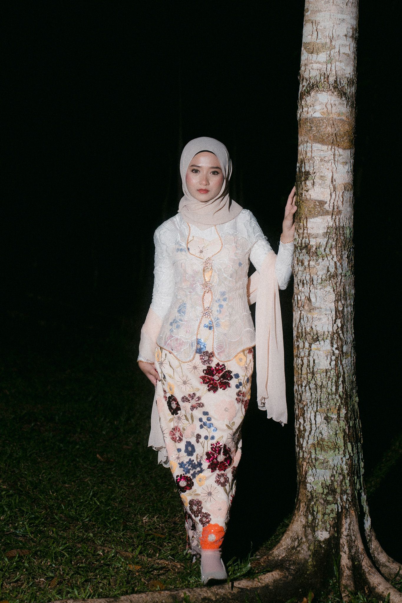 Woman in white hijab and floral kebaya skirt poses by a tree at night.