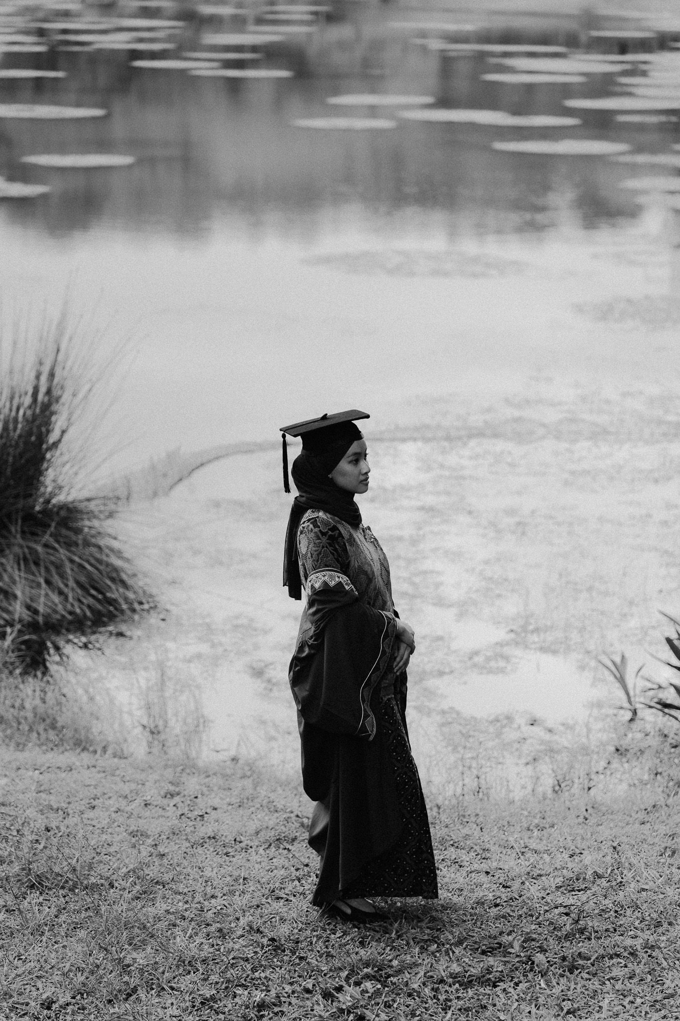 Black and white photo of a graduate in cap and gown standing by a serene pond with lily pads.
