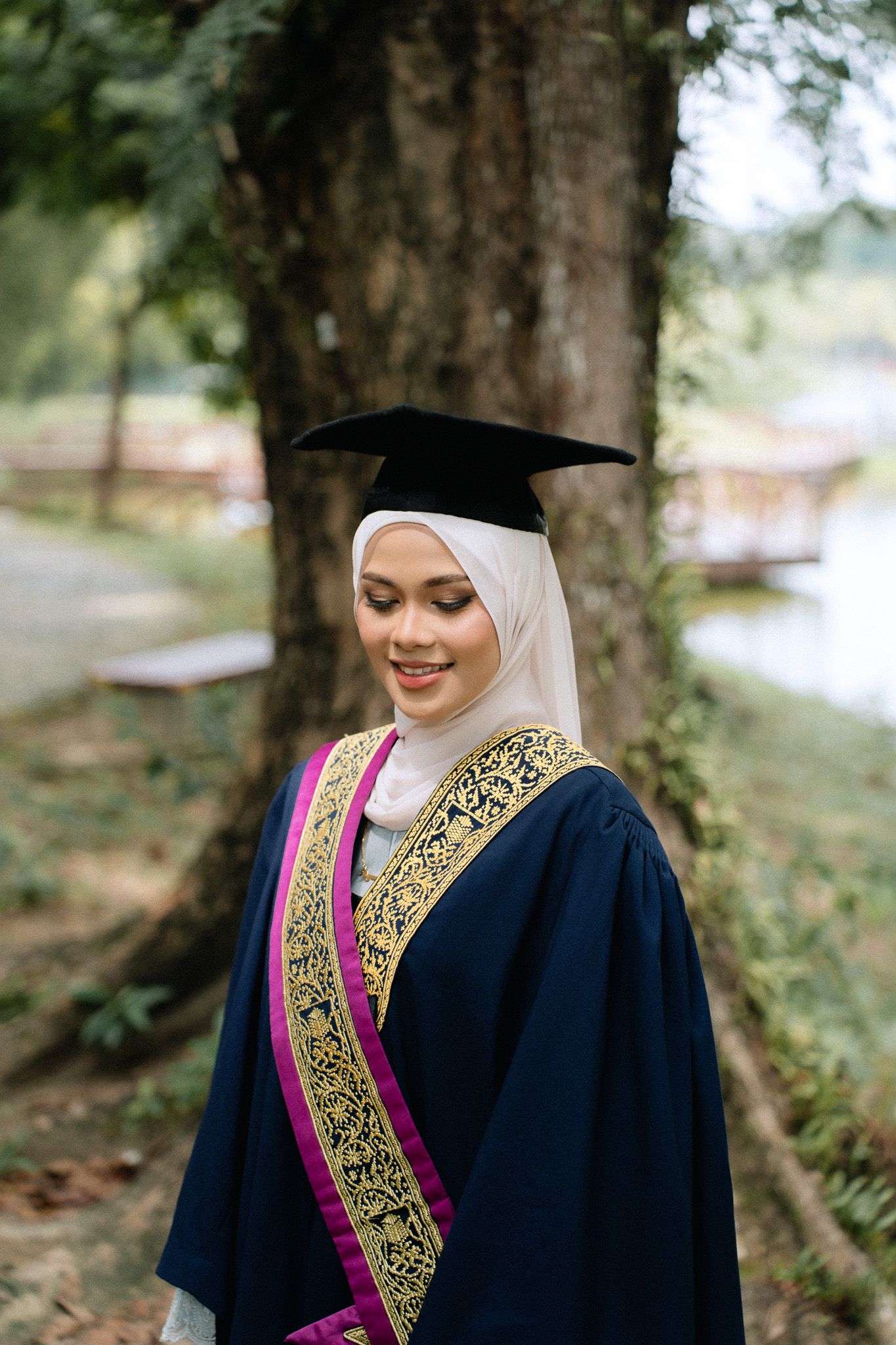Muslim woman in hijab and graduation gown with mortarboard cap smiling outdoors near a tree.