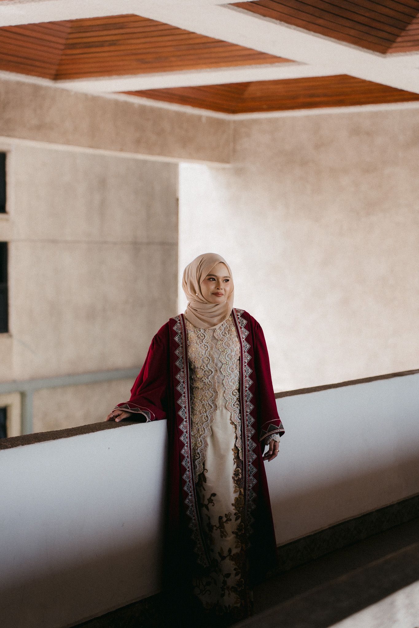 Woman in burgundy embroidered abaya and beige hijab standing on a balcony in elegant traditional Muslim attire.