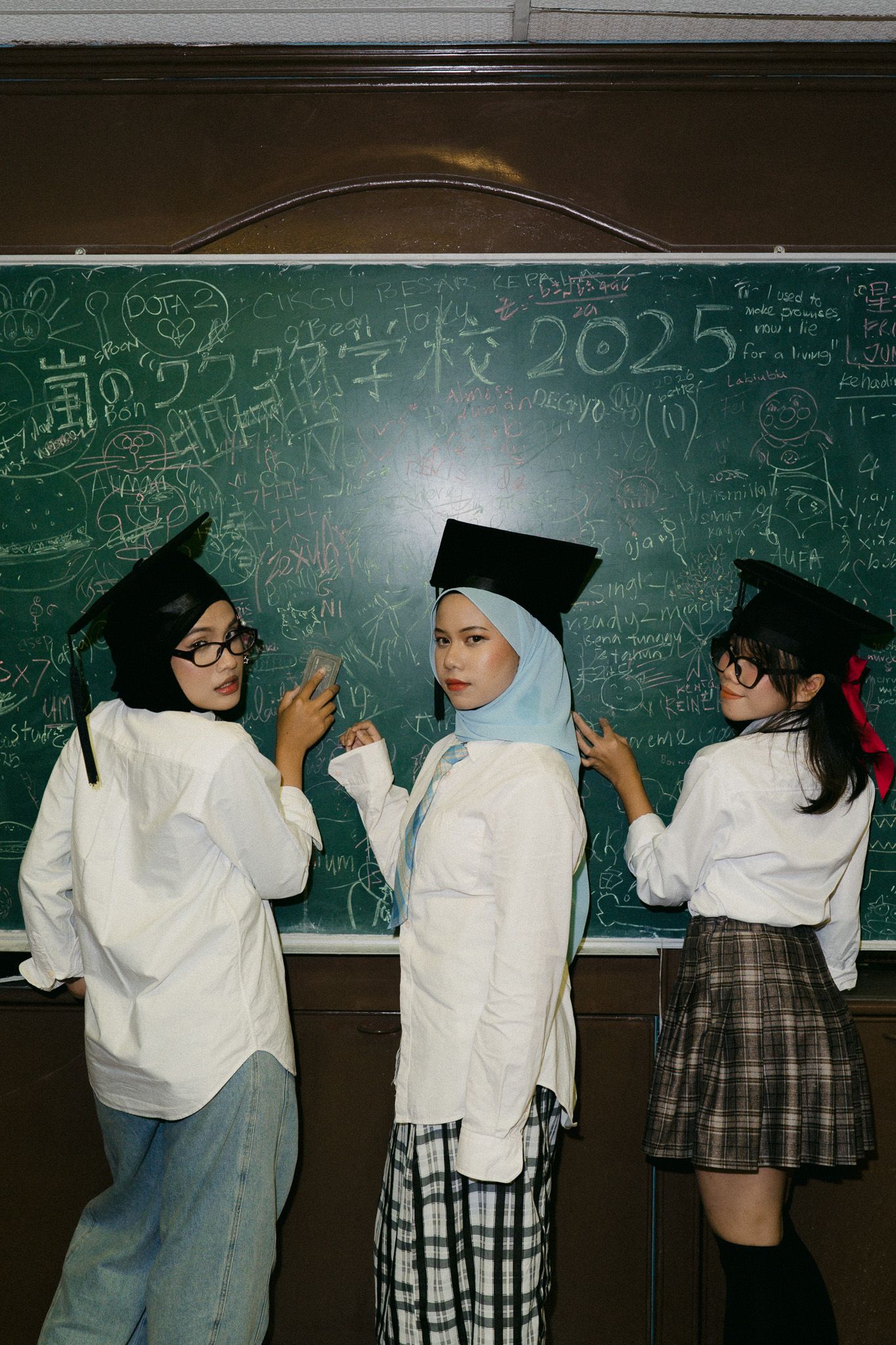 Three students in graduation caps and white shirts pose together in front of a green chalkboard in a classroom.