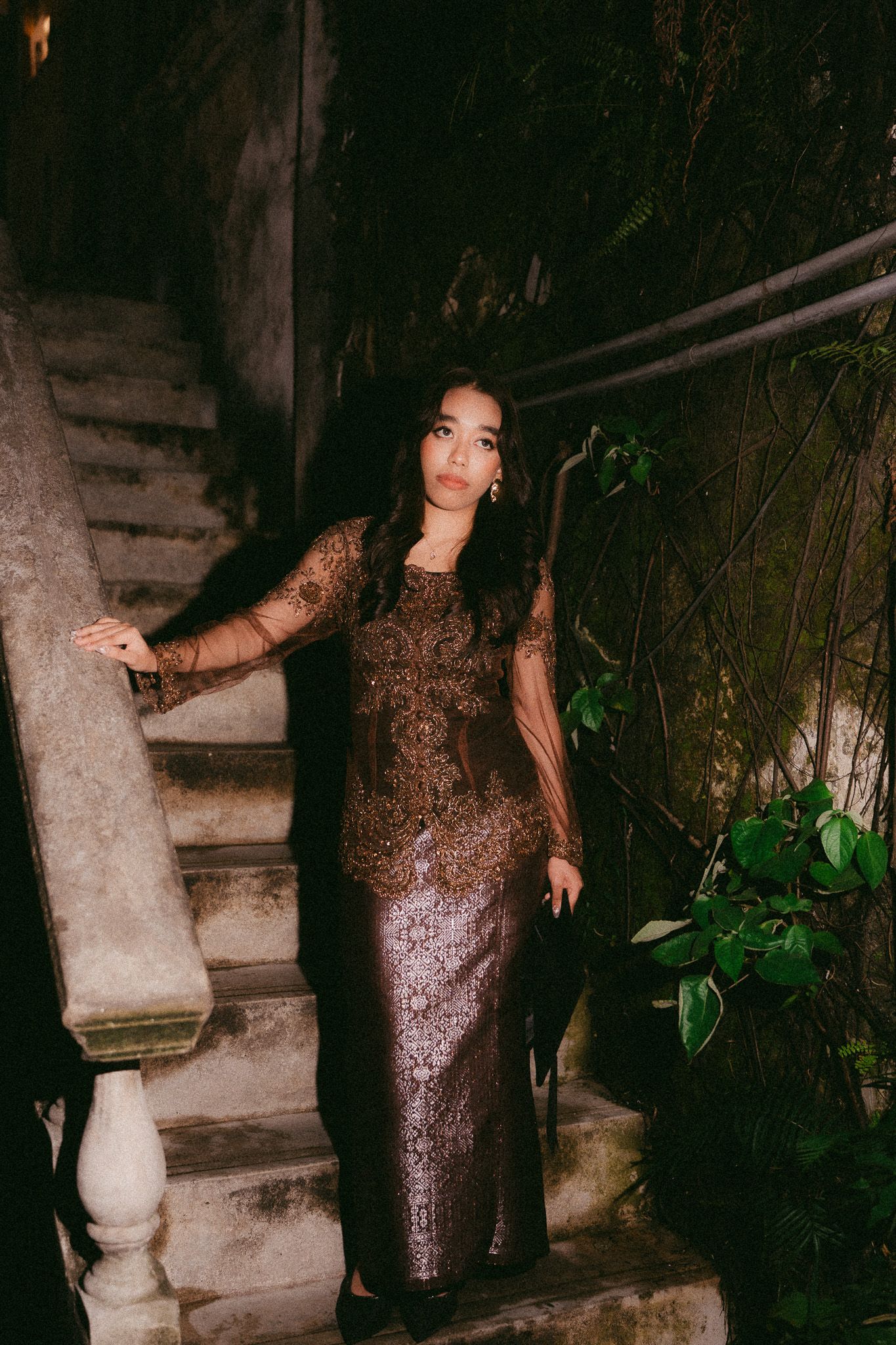 Woman in elegant gold embroidered gown poses on stone staircase surrounded by lush greenery at night.
