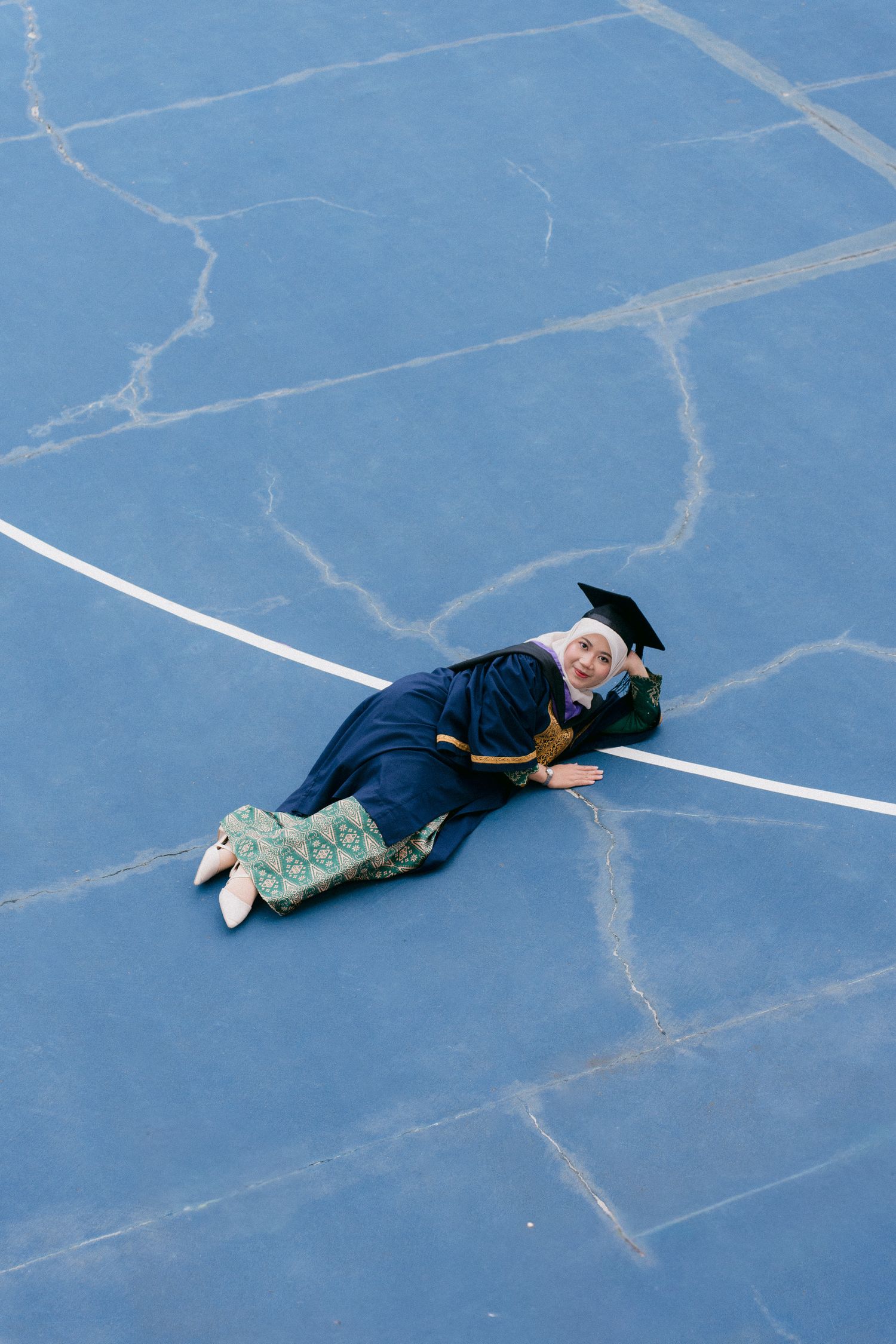 Graduate in cap and gown lies on blue tennis court during celebratory photo session.
