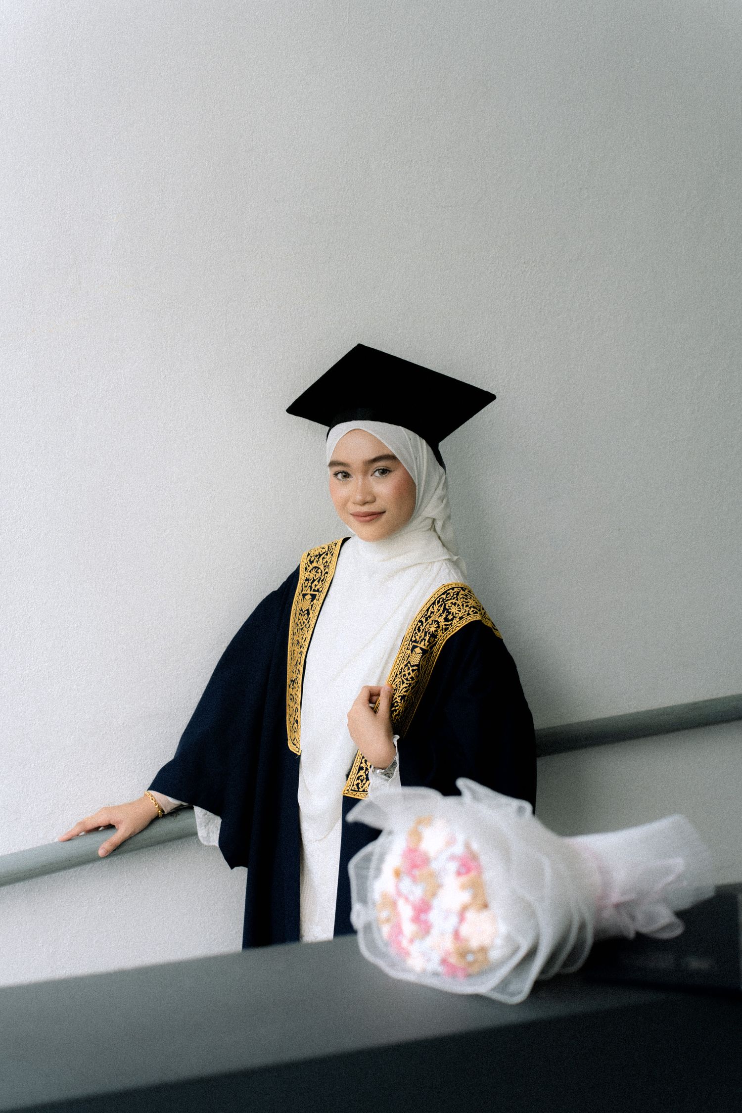Graduate in black academic regalia and white hijab poses playfully in a sequence of artistic photos against grey wall.
