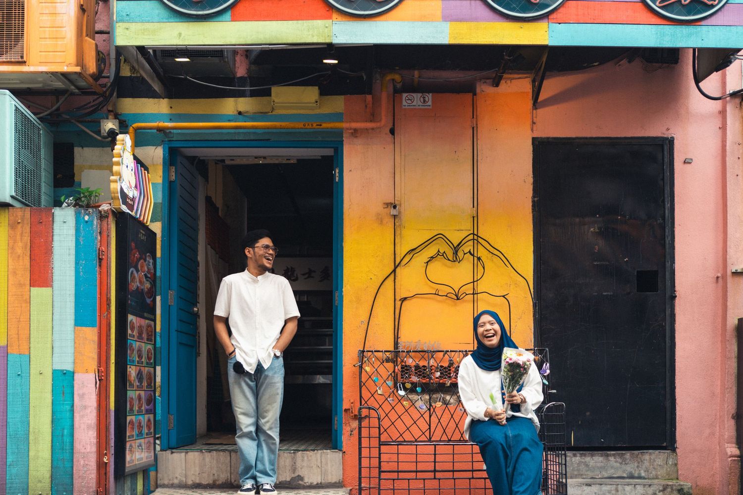 Two people stand against colorful painted walls and doorways in an urban setting.