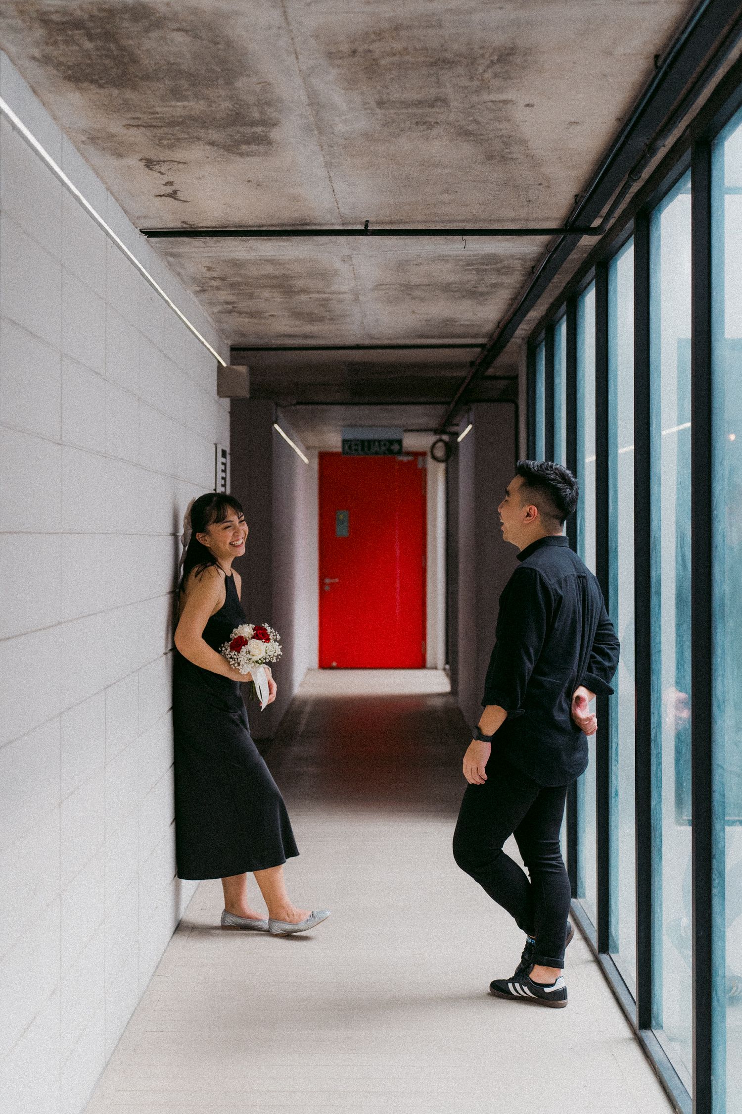 Two people standing in a minimalist hallway with white walls and a striking red door at the end.