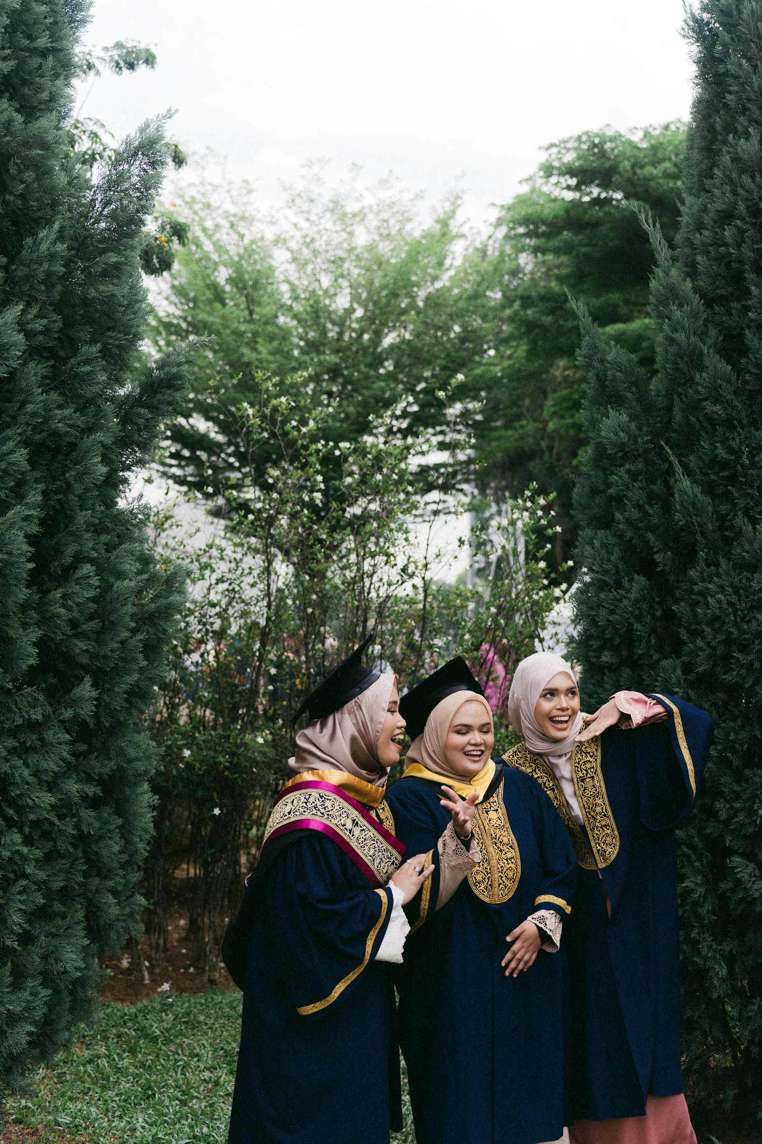 Three graduates in navy blue robes with gold trim and headscarves share a joyful moment together in a garden setting.