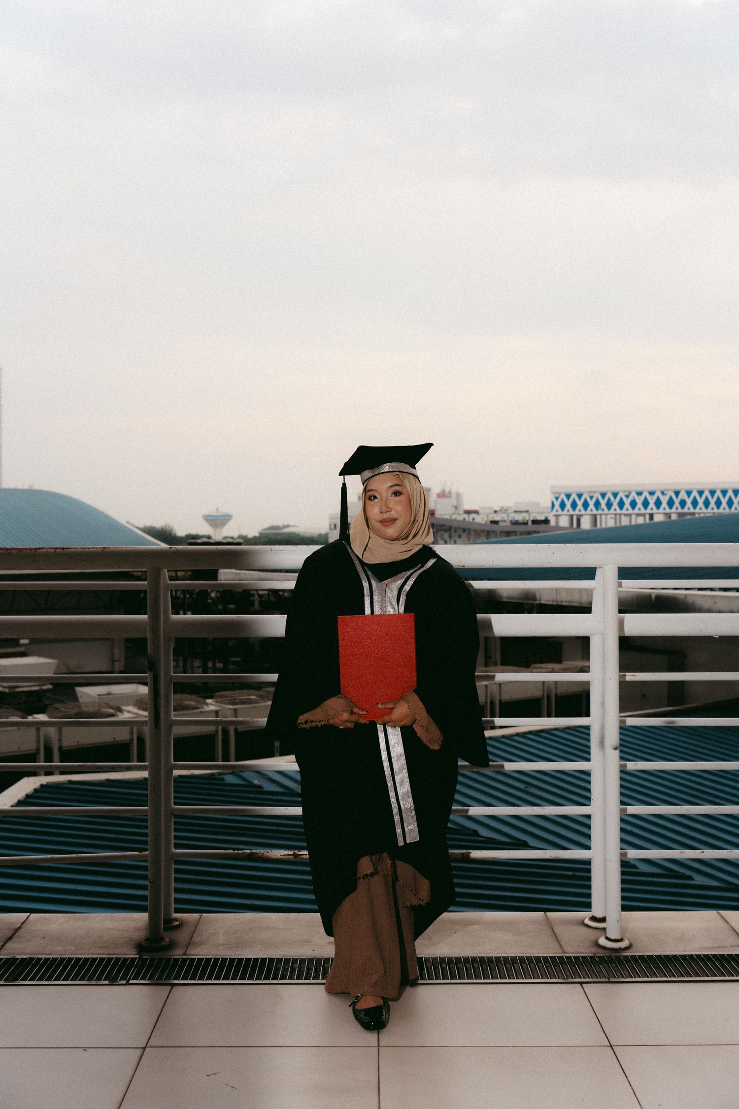 A graduate wearing a black cap and gown holds a red diploma folder while sitting on a railing outdoors.