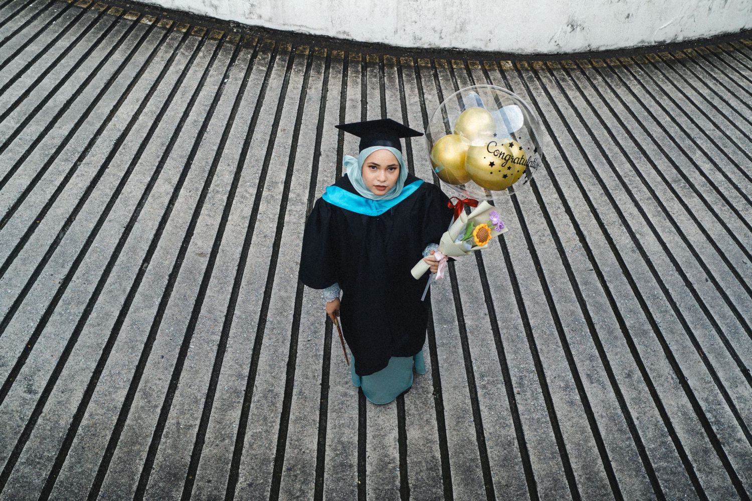Graduate in black gown holds gold balloons while standing on textured surface.