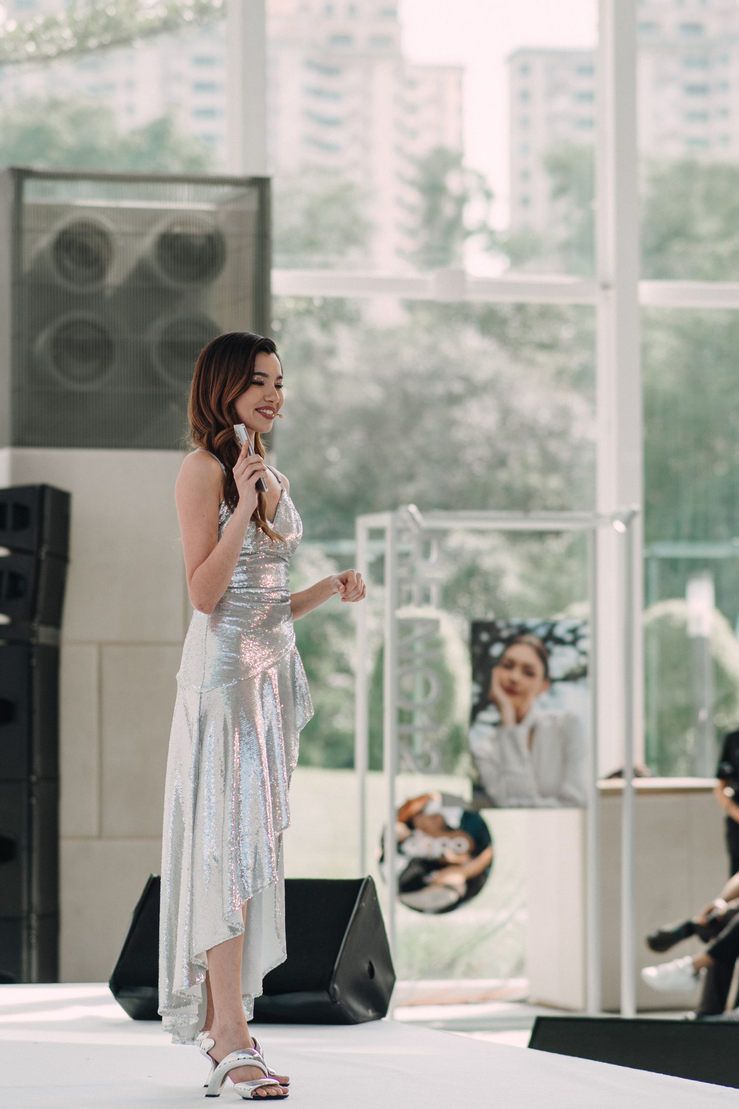 Fashion model poses in shimmering silver dress on stage during product launch event.