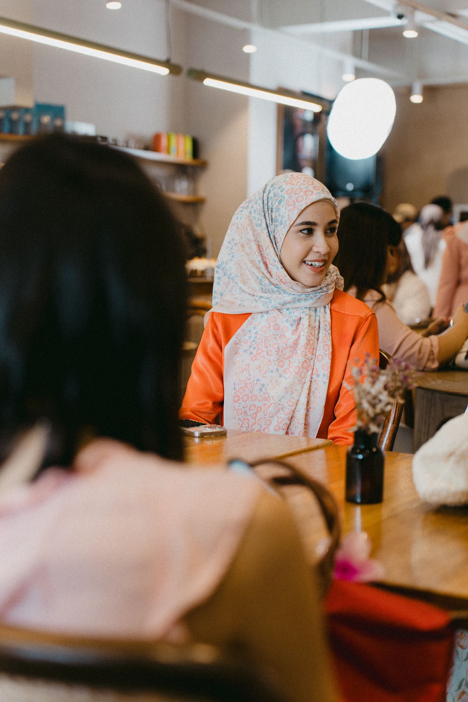 A woman wearing an orange top and floral hijab smiles while seated at a cafe counter.