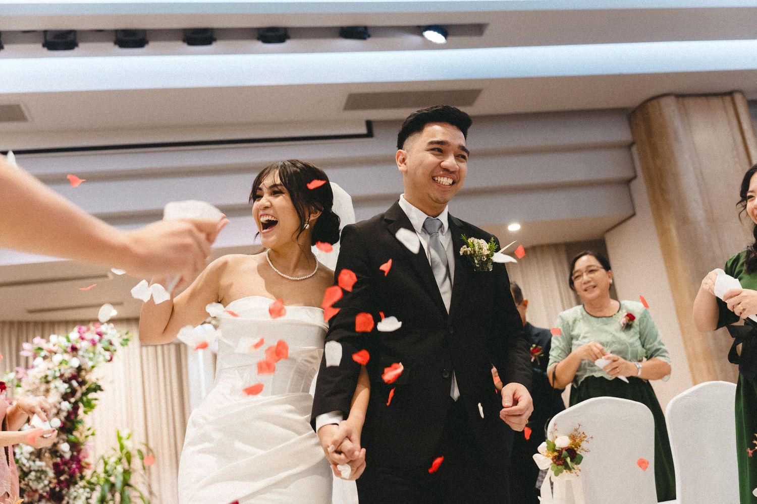 Joyful wedding ceremony with rose petals being thrown as couple walks down decorated indoor aisle.