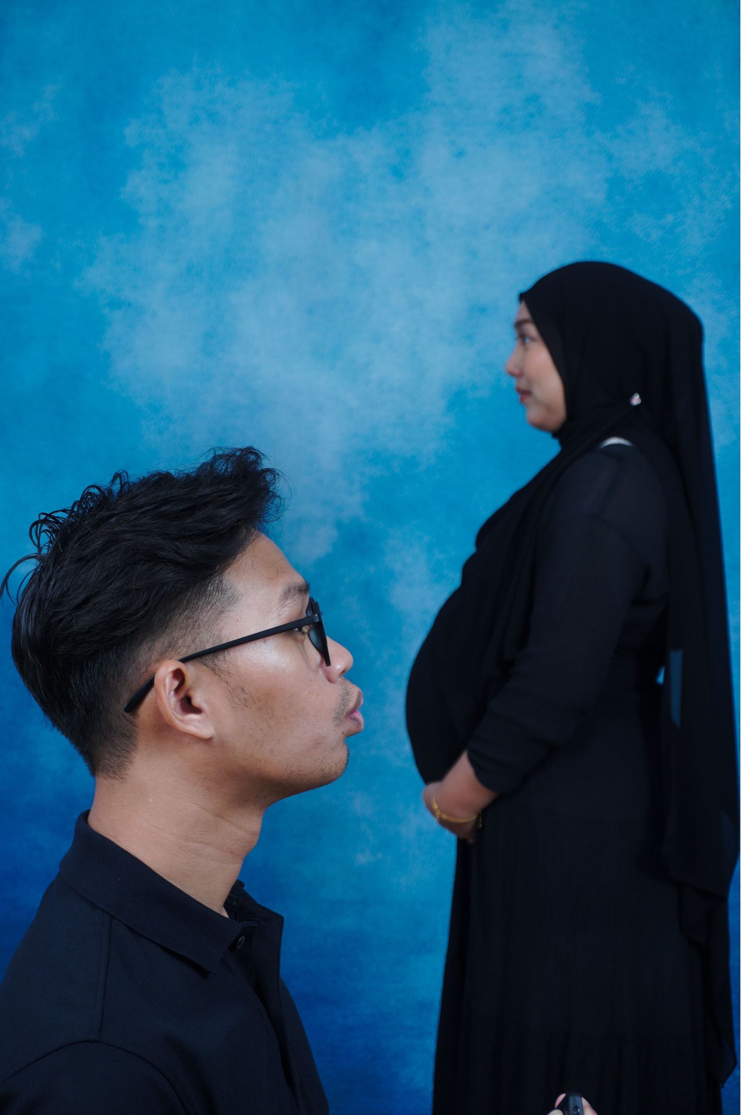 A man in glasses and a pregnant woman in black hijab pose against a blue backdrop.