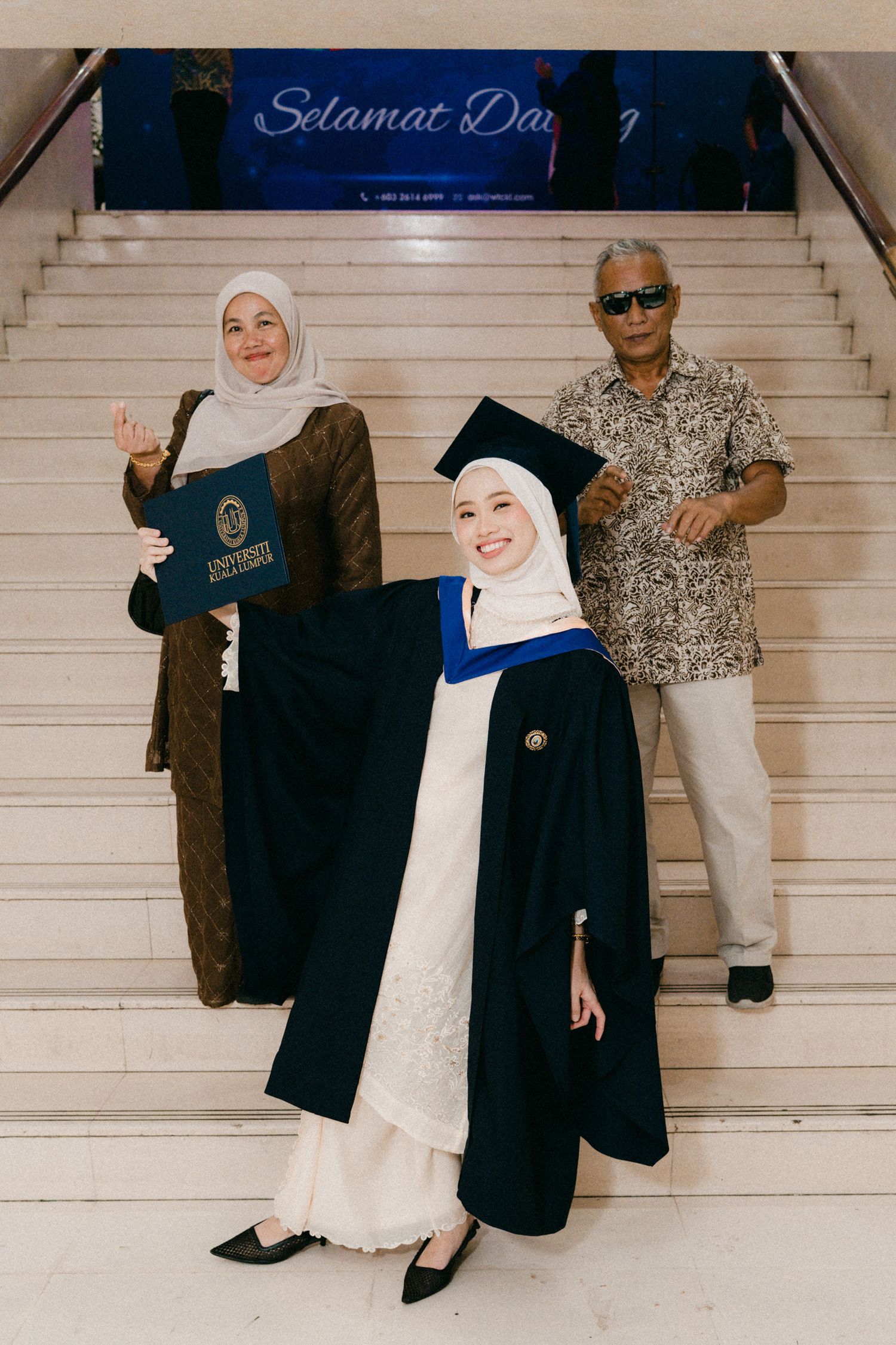 Graduation celebration photo on stairs with graduate in black academic regalia, white dress and hijab under a blue banner.
