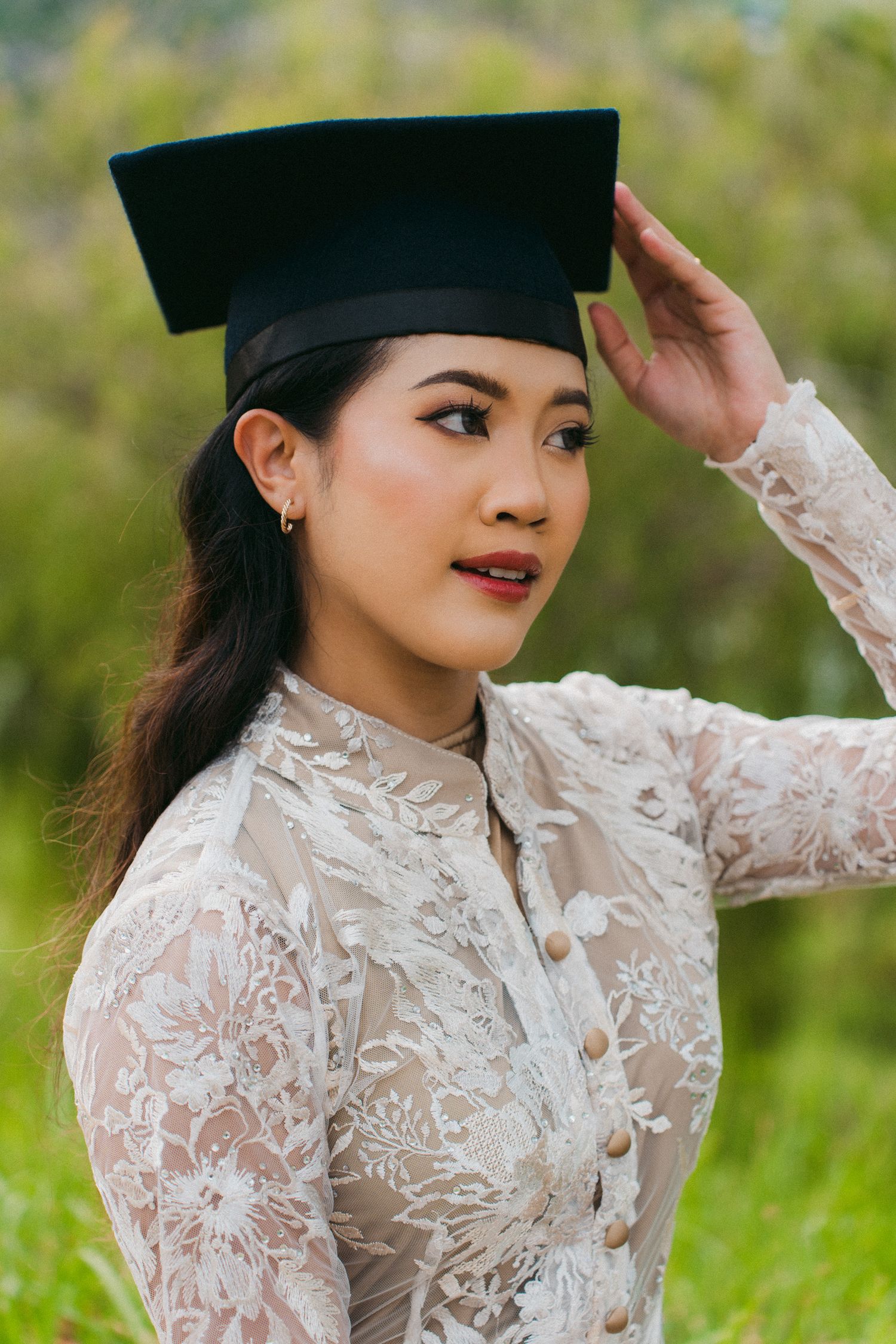 Graduate in white lace traditional dress and academic cap poses in outdoor garden setting with natural greenery.