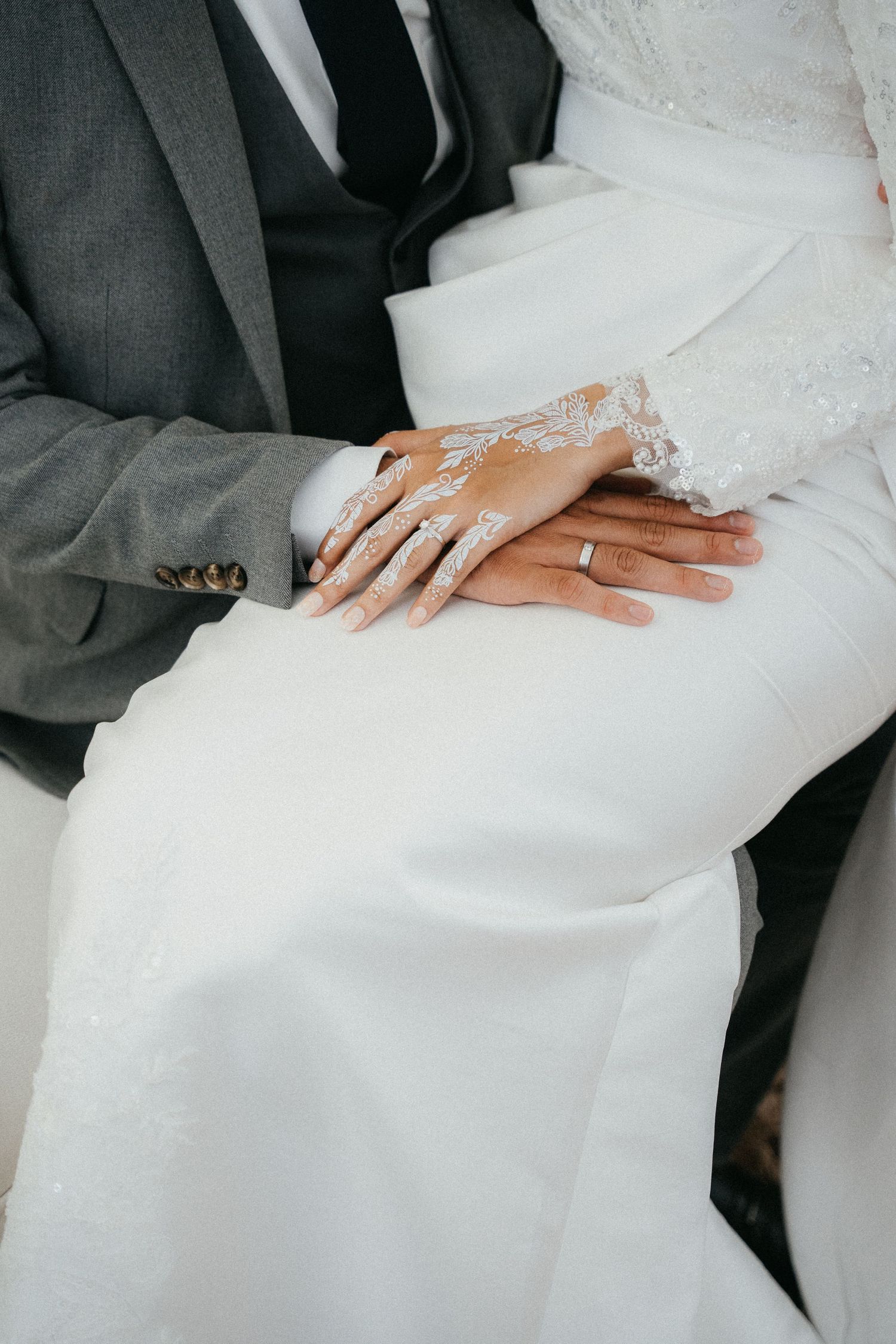 Close-up of newlywed couple's hands with wedding rings resting on white wedding dress and gray suit.
