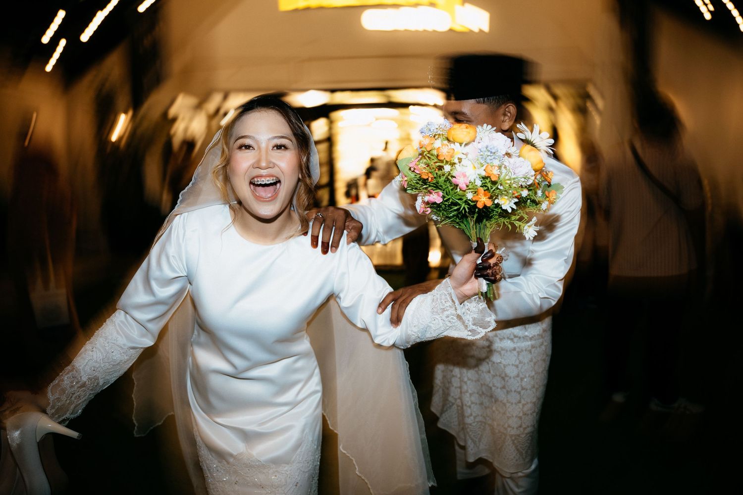 Joyful couple running down a dimly lit hallway with a bouquet of flowers after their wedding celebration.
