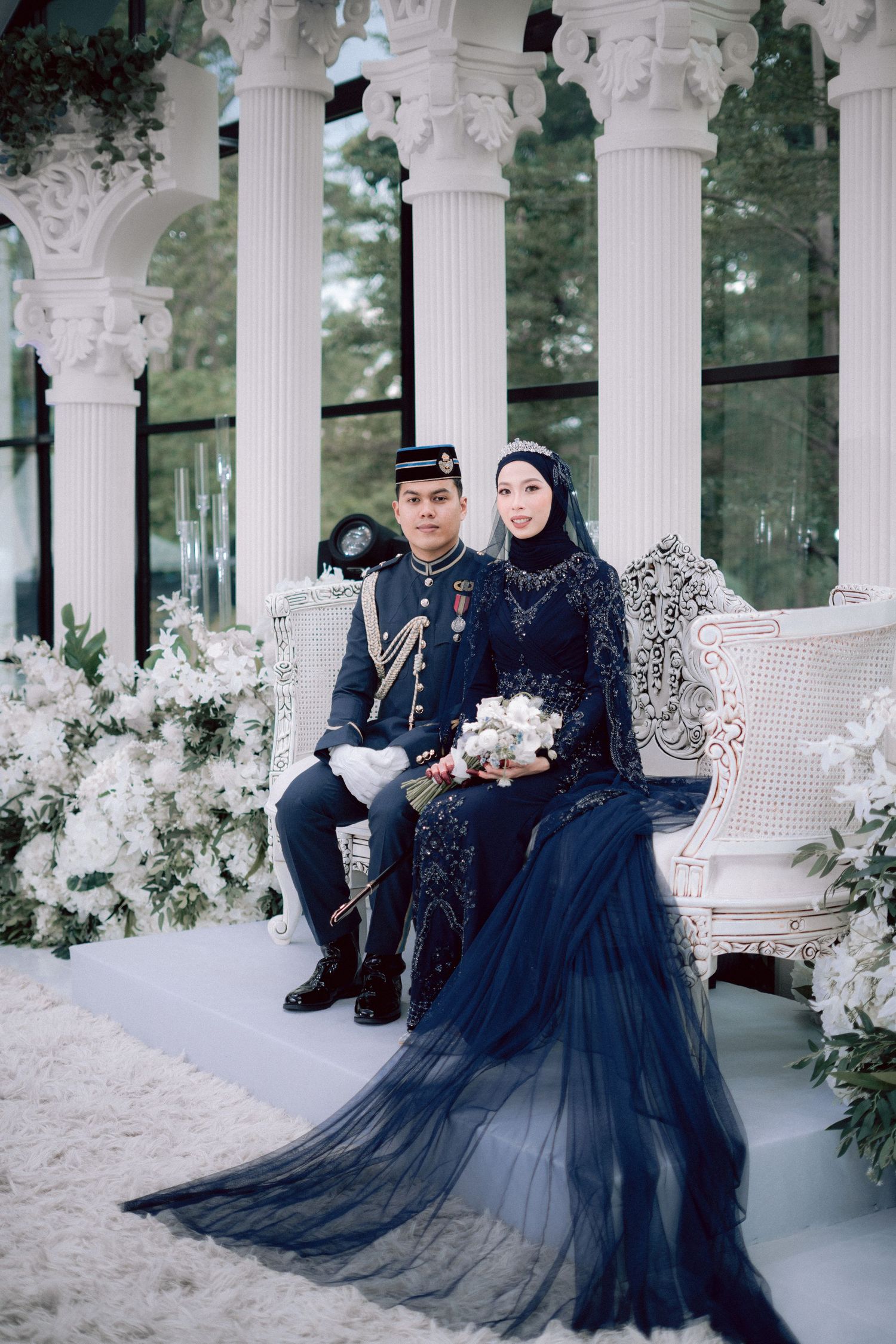 A couple in formal attire sits on an elegant white columned porch decorated with winter flowers and white decor.