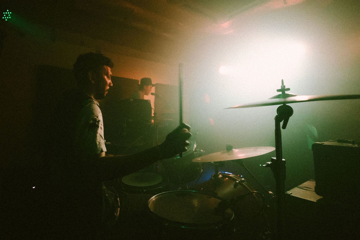 A drummer plays behind a drum kit on stage with atmospheric green lighting and fog in the background.