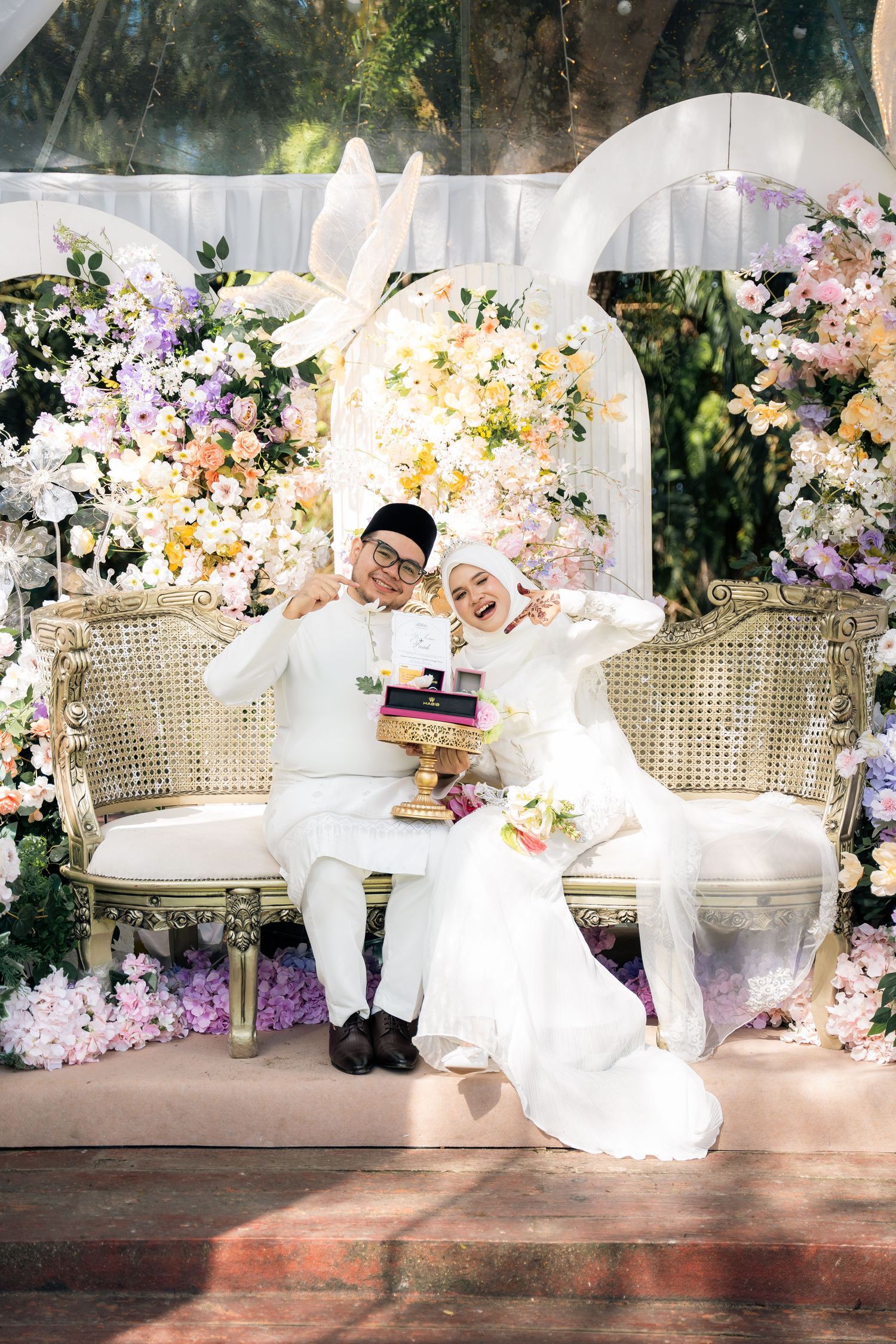 A romantic scene with couple in white attire on an elegant settee surrounded by lush floral arrangements.