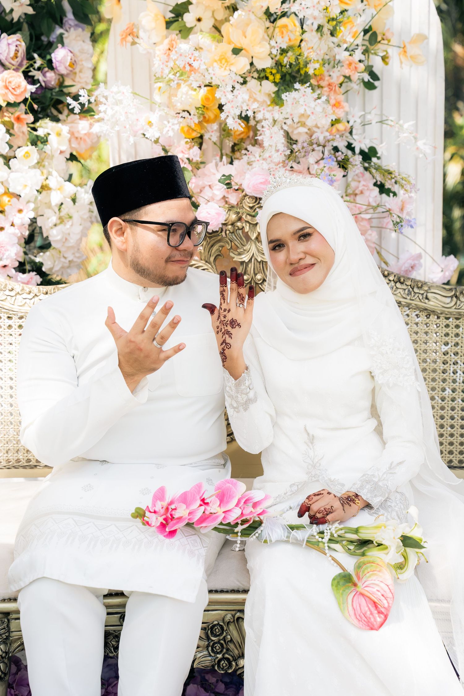 Traditional Malay wedding couple in white attire seated on ornate bench with floral backdrop of pink and yellow blooms.