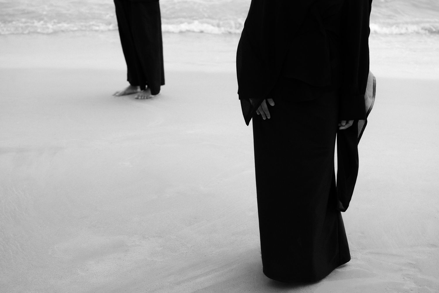 Two figures in black attire stand on a sandy beach in a moody black and white photograph.