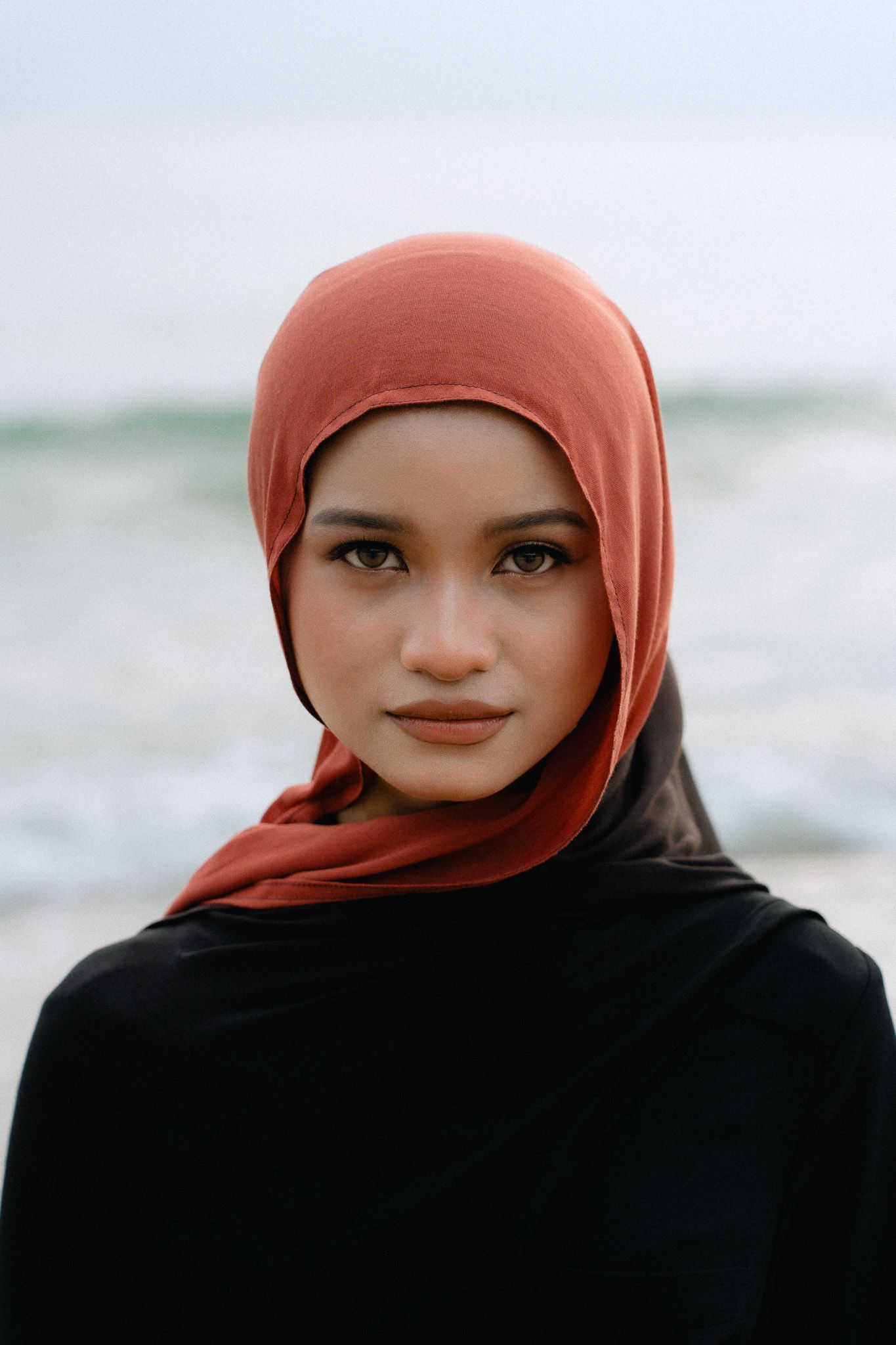 Young woman wearing a rust-colored hijab and black outfit posing on a beach with ocean waves behind her.