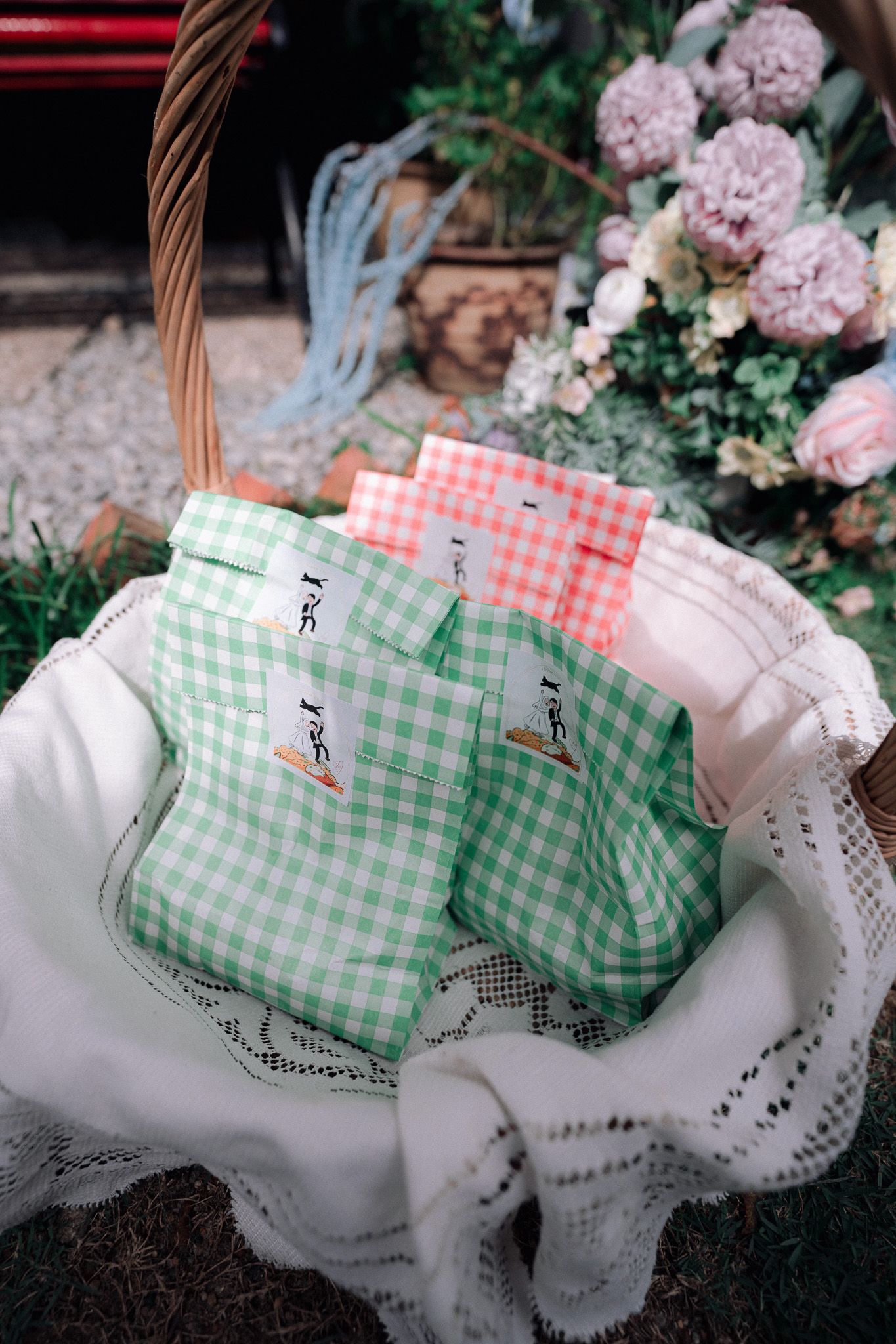 Green and pink gingham gift bags in a wicker basket lined with white lace cloth, surrounded by fresh flowers.
