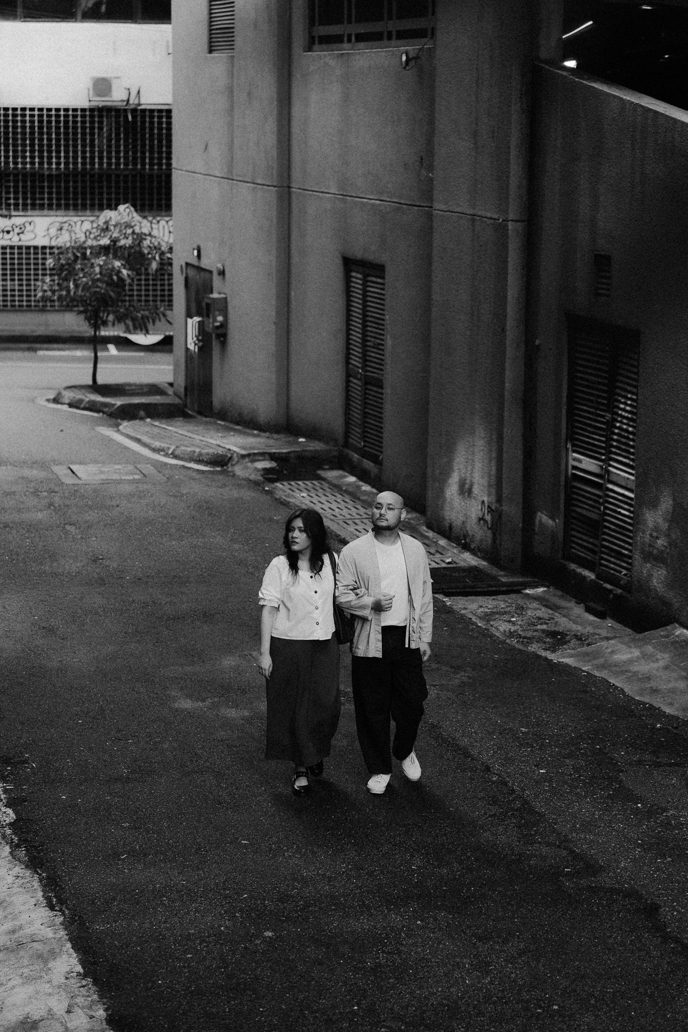 Black and white photo of a couple walking together on an urban street beside a concrete building.