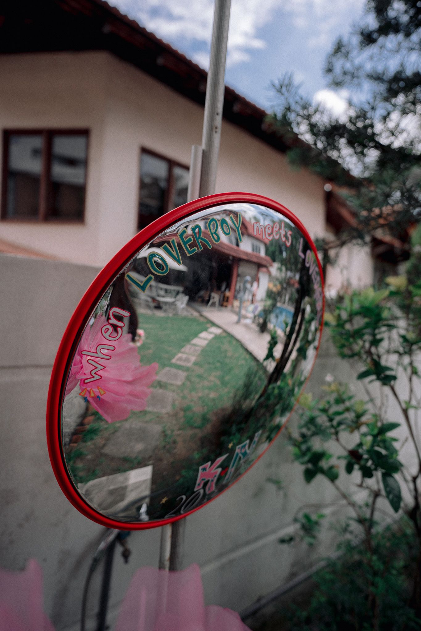 Convex safety mirror with red frame mounted on a pole, reflecting a colorful storefront and green garden.