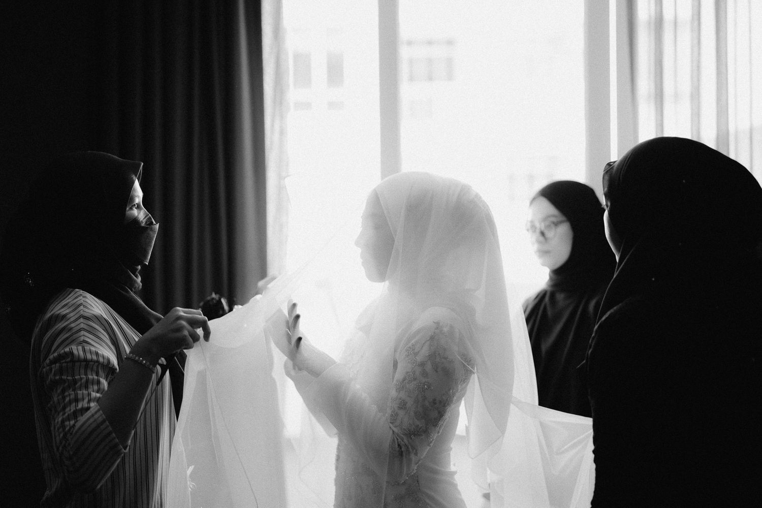 Black and white photo of a bride in a veil being assisted by women during wedding preparations near a bright window.