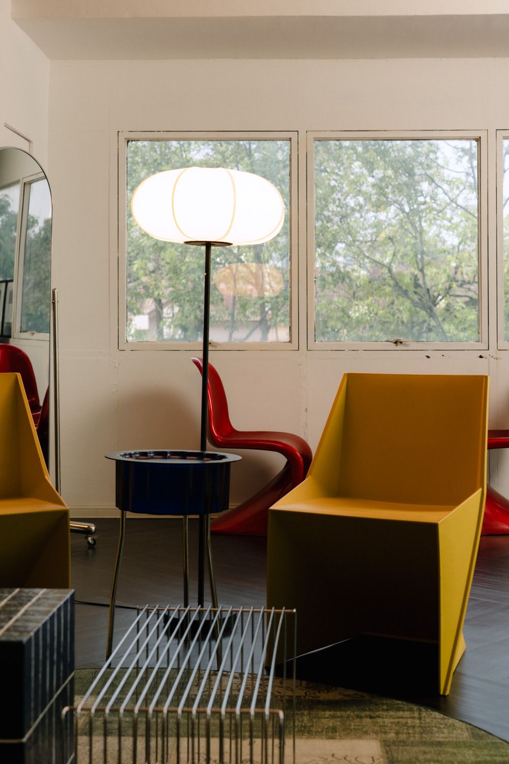 Modern living room with yellow chairs, red accent chair, white floor lamp, and large windows overlooking green trees.