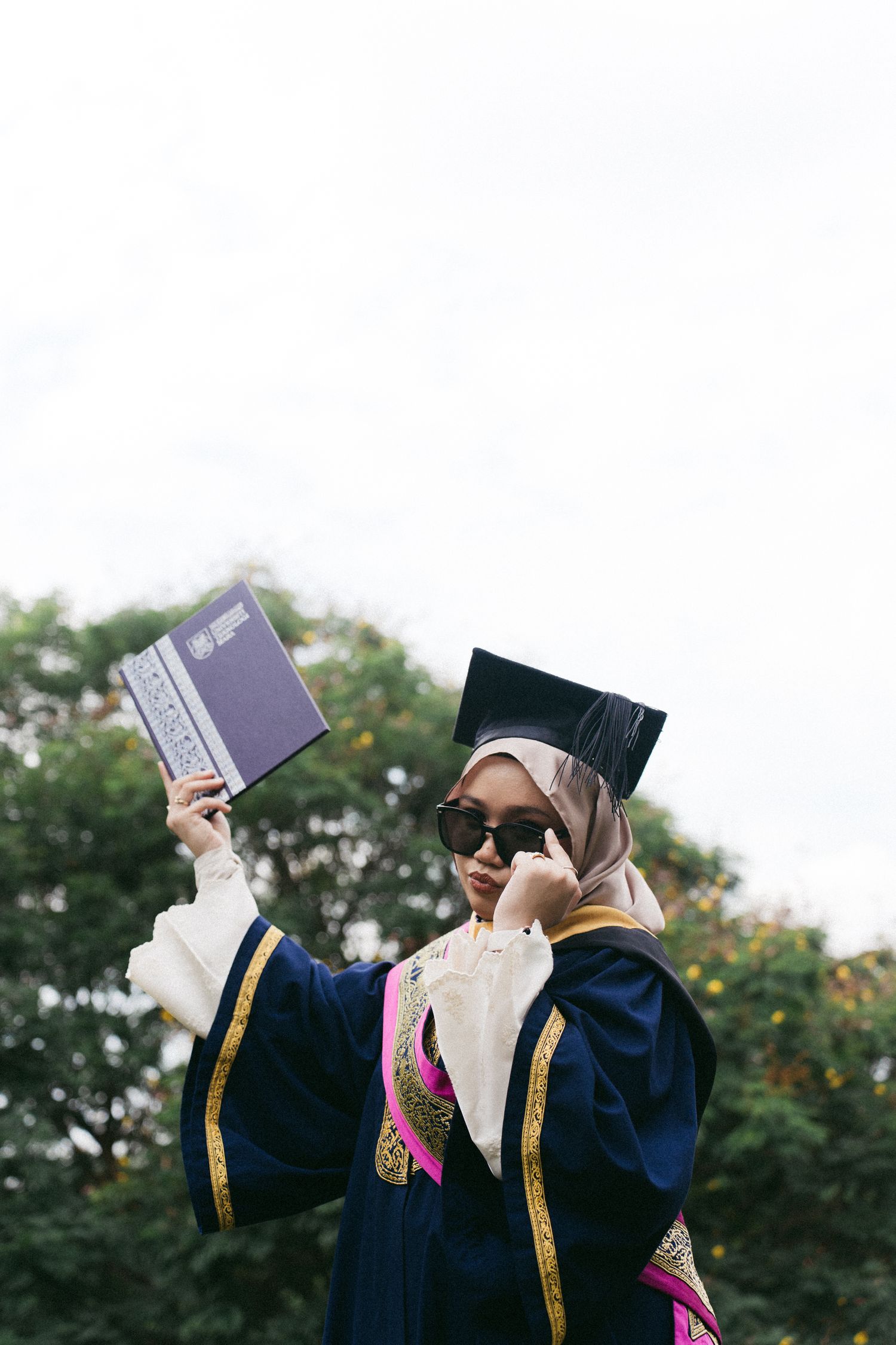 Graduate in blue academic regalia and hijab holds up diploma and celebrates during outdoor ceremony.