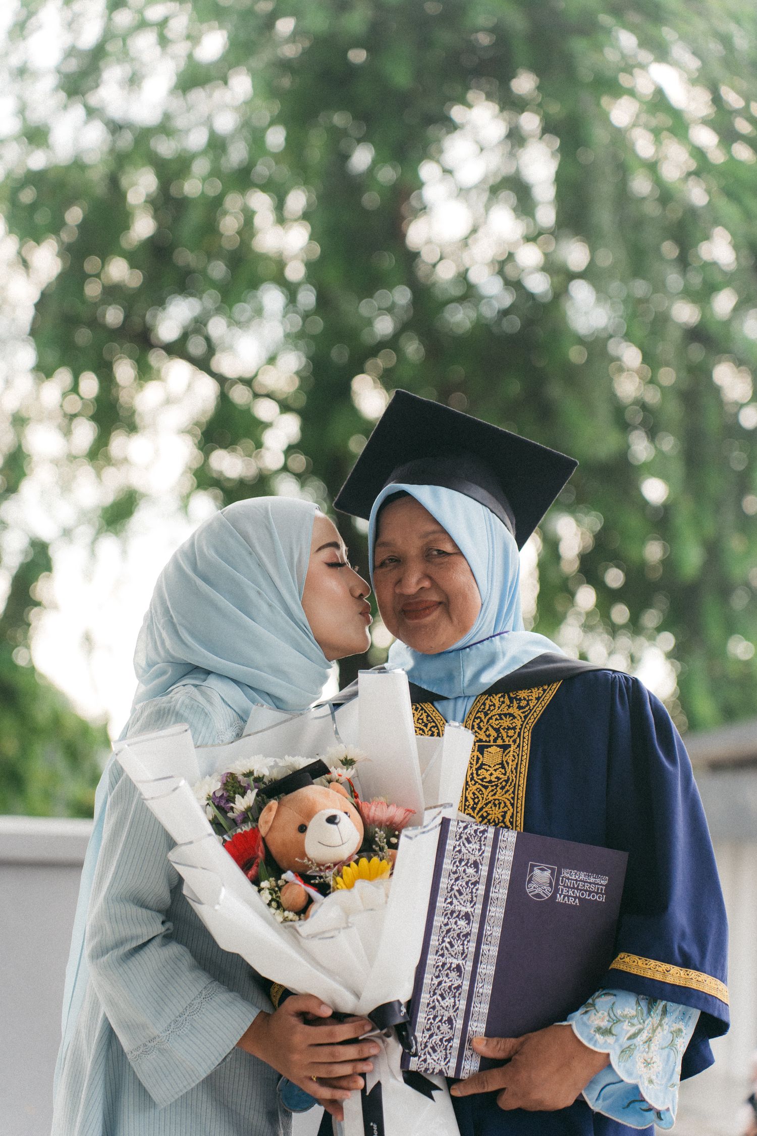 Two women in light blue hijabs share a tender moment at graduation, with one holding a bouquet and teddy bear.