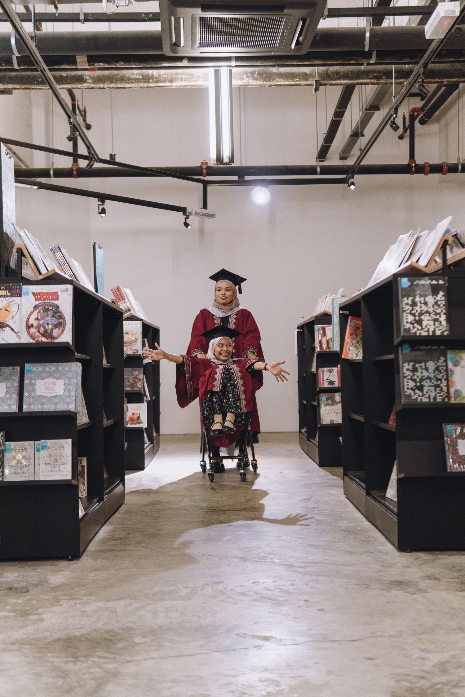A graduate in red academic regalia stands between rows of bookshelves in a library.