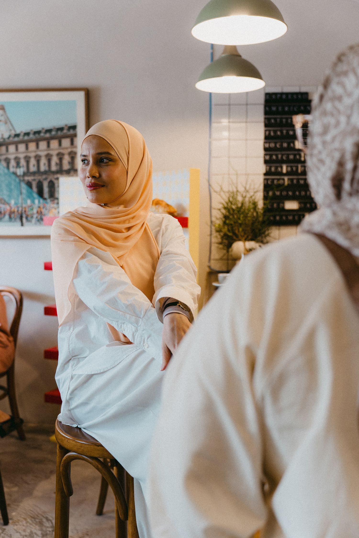 Person in white uniform and hijab seated in modern cafe interior.