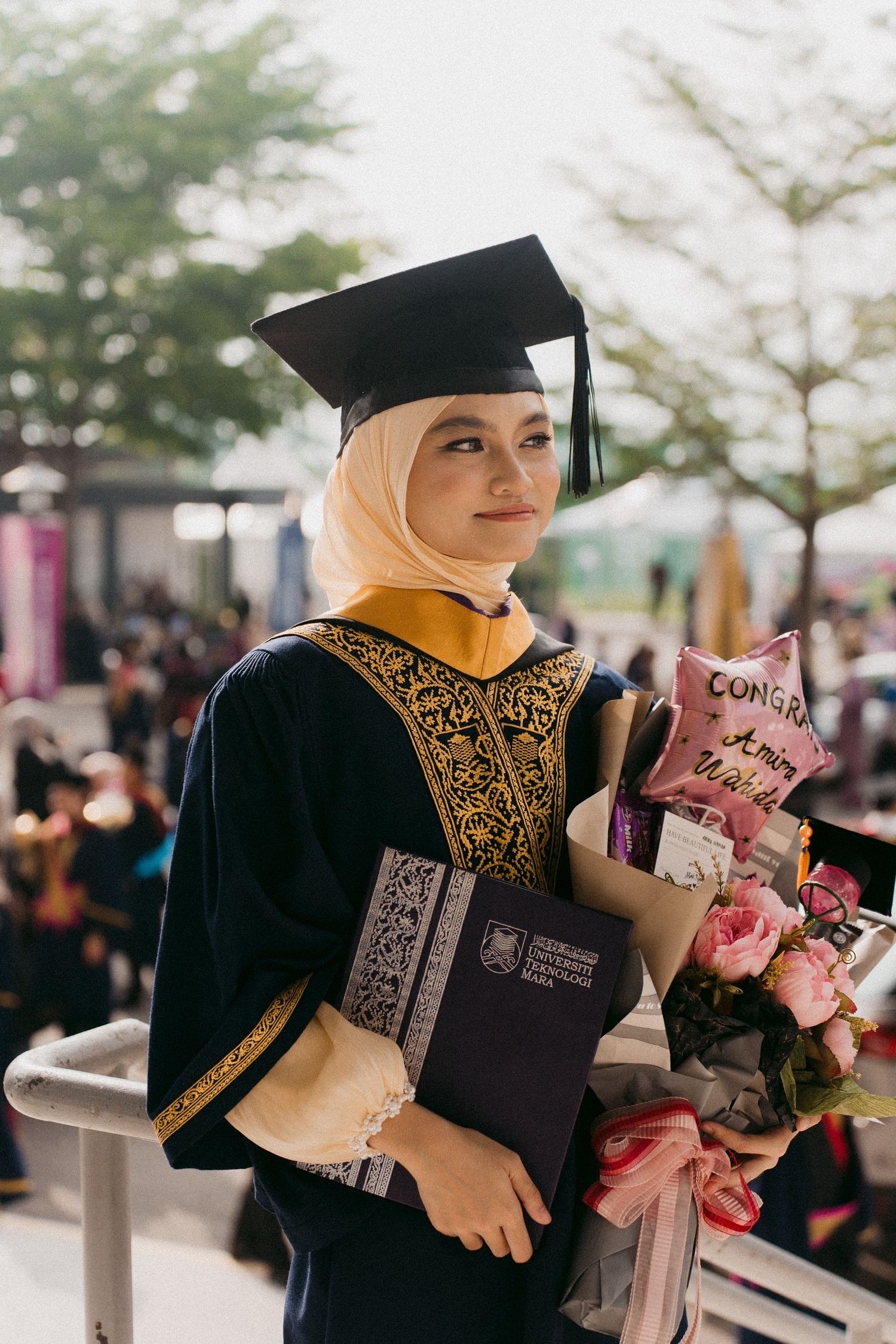 Graduate in black and gold academic regalia holds flowers and diploma during outdoor graduation ceremony.
