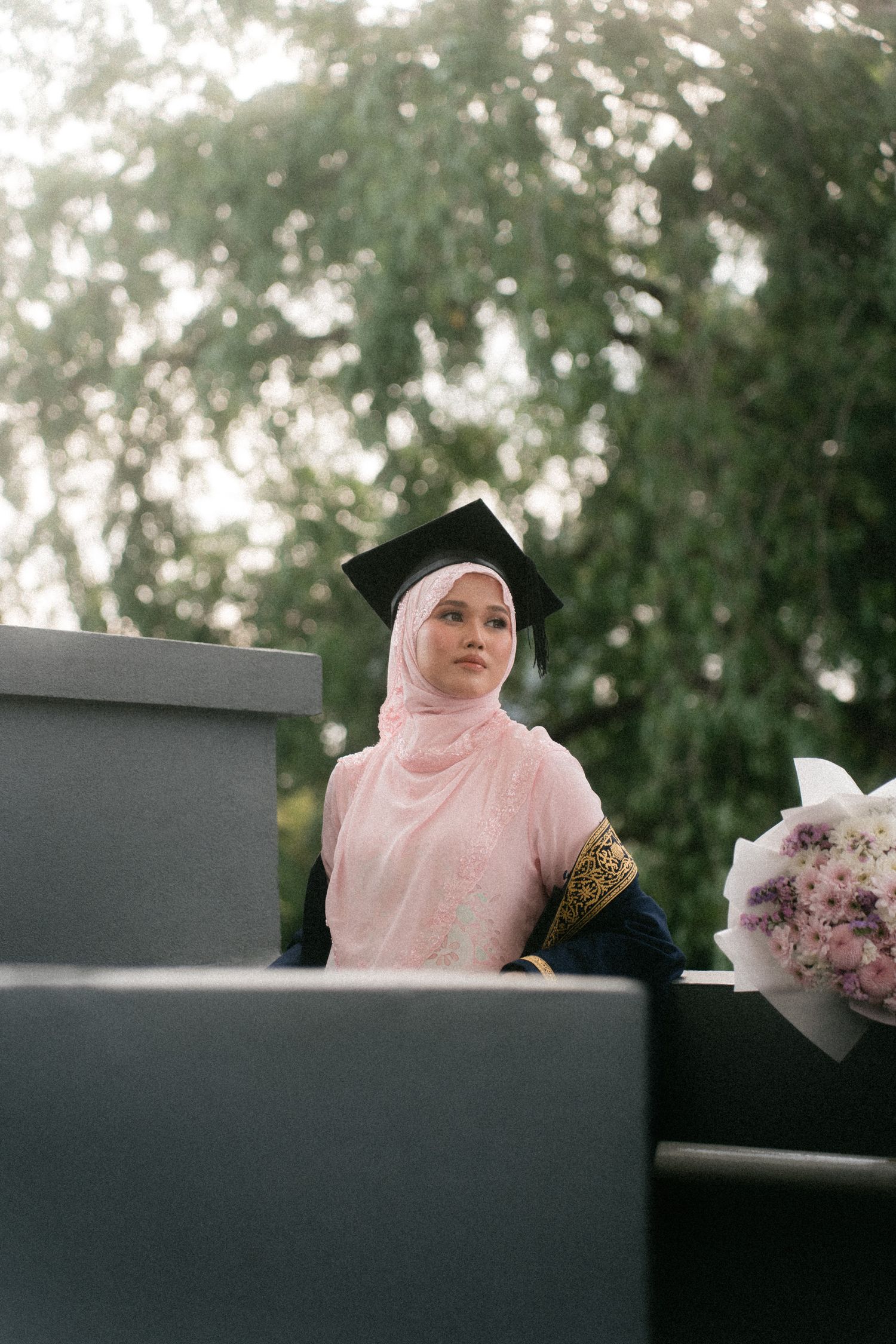 A person in a pink hijab sits at a dark desk with flowers in an outdoor garden setting.