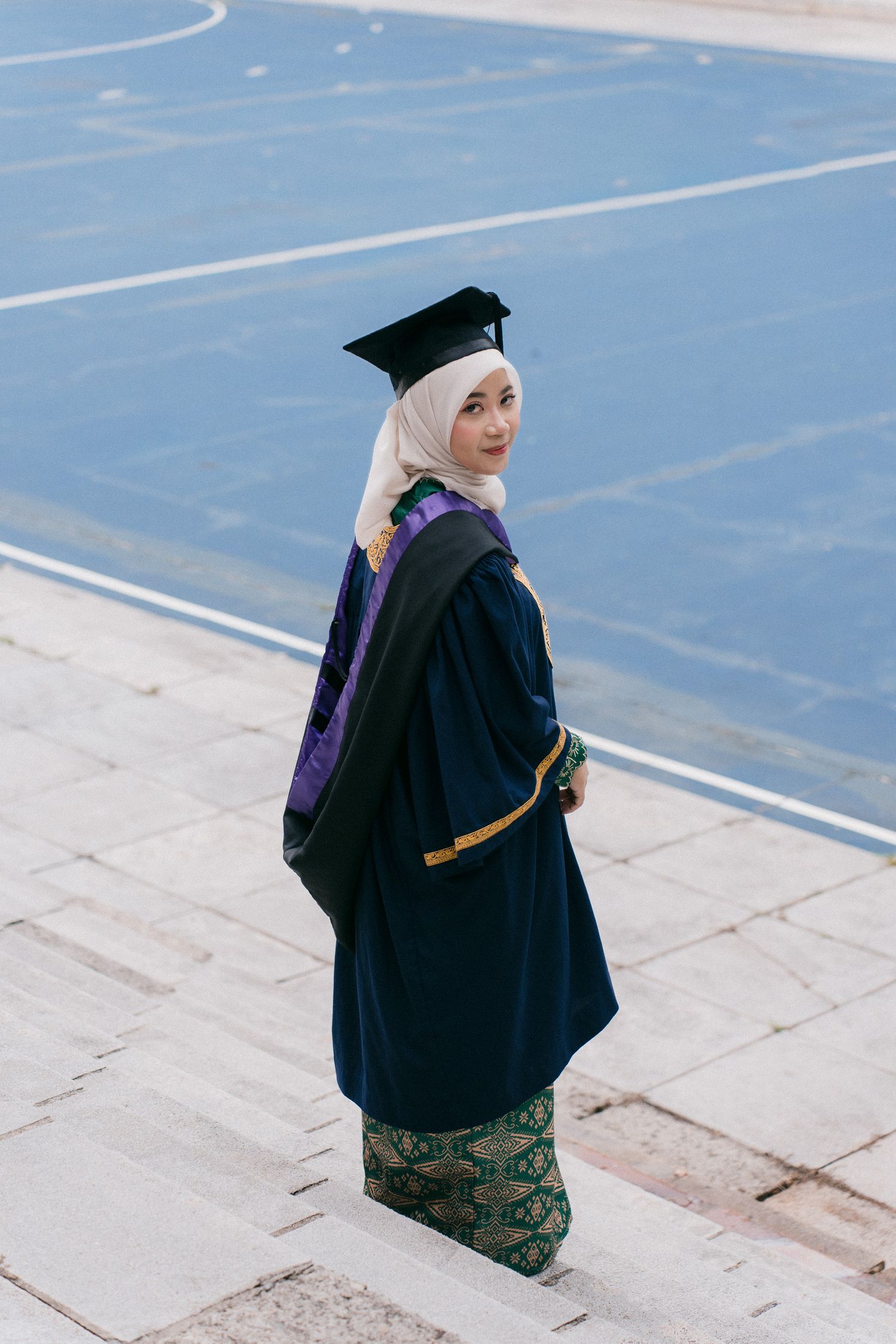 Graduate in hijab and academic regalia walking on blue tiled surface during commencement ceremony.