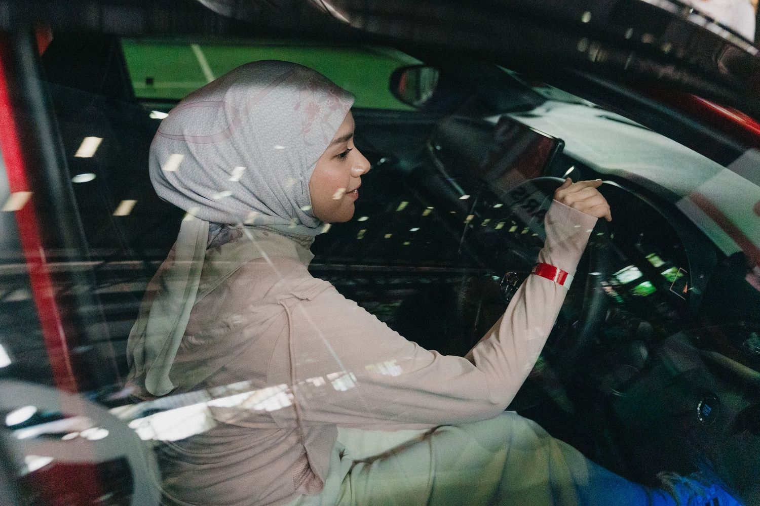 A person wearing a light beige hijab sits in the driver's seat of a car, viewed through the side window.
