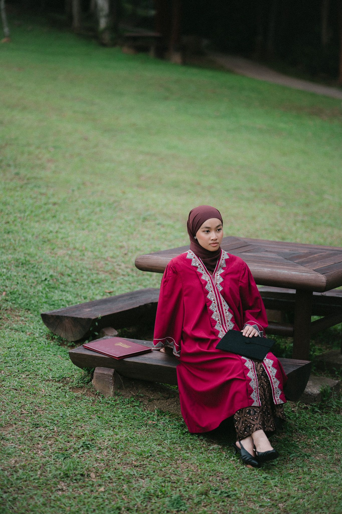A young Muslim woman in a red hijab and embroidered dress sits on a wooden bench in a lush green park.