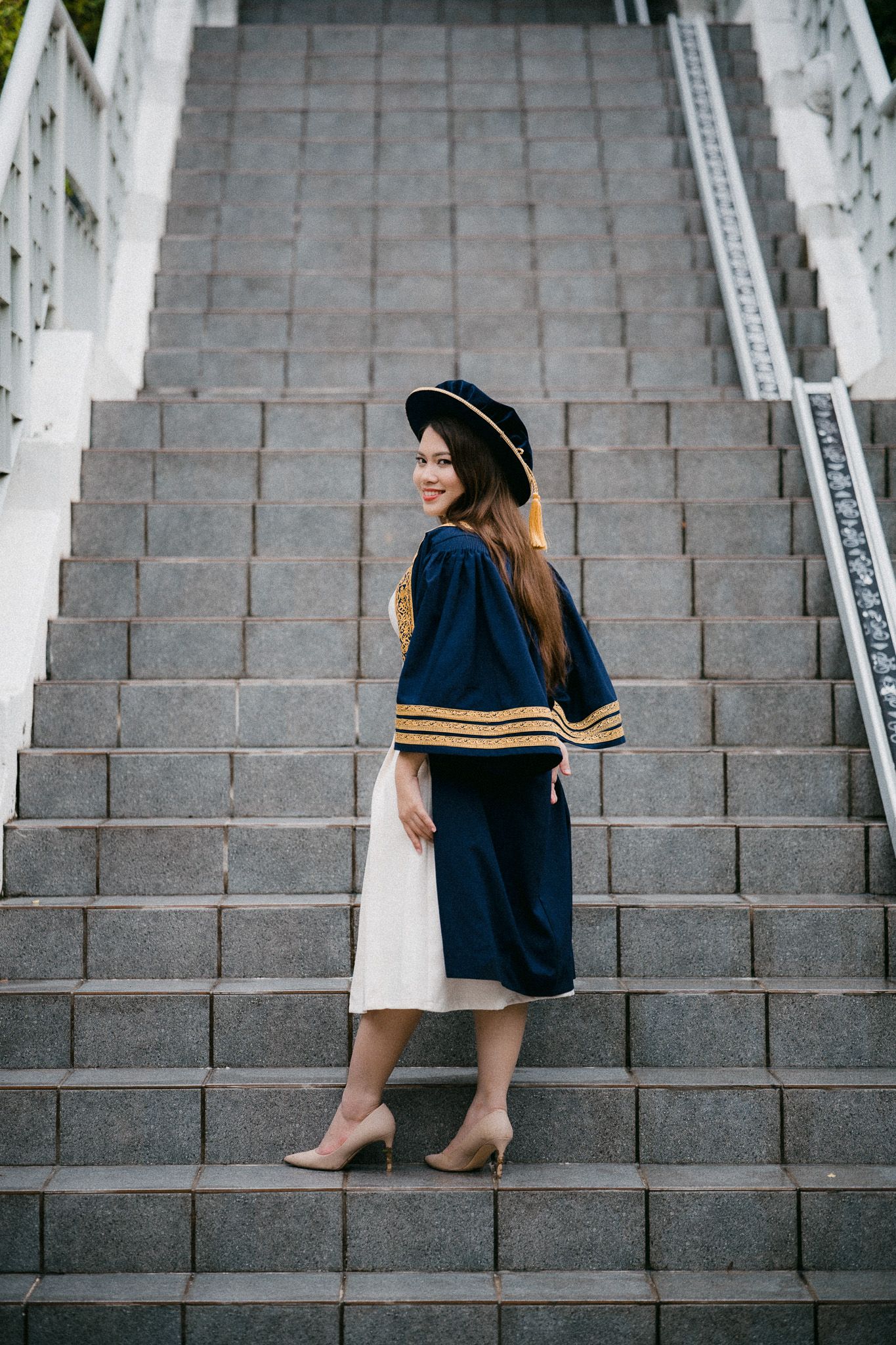 Young woman in graduation gown and black hat poses on stone staircase, smiling over her shoulder.