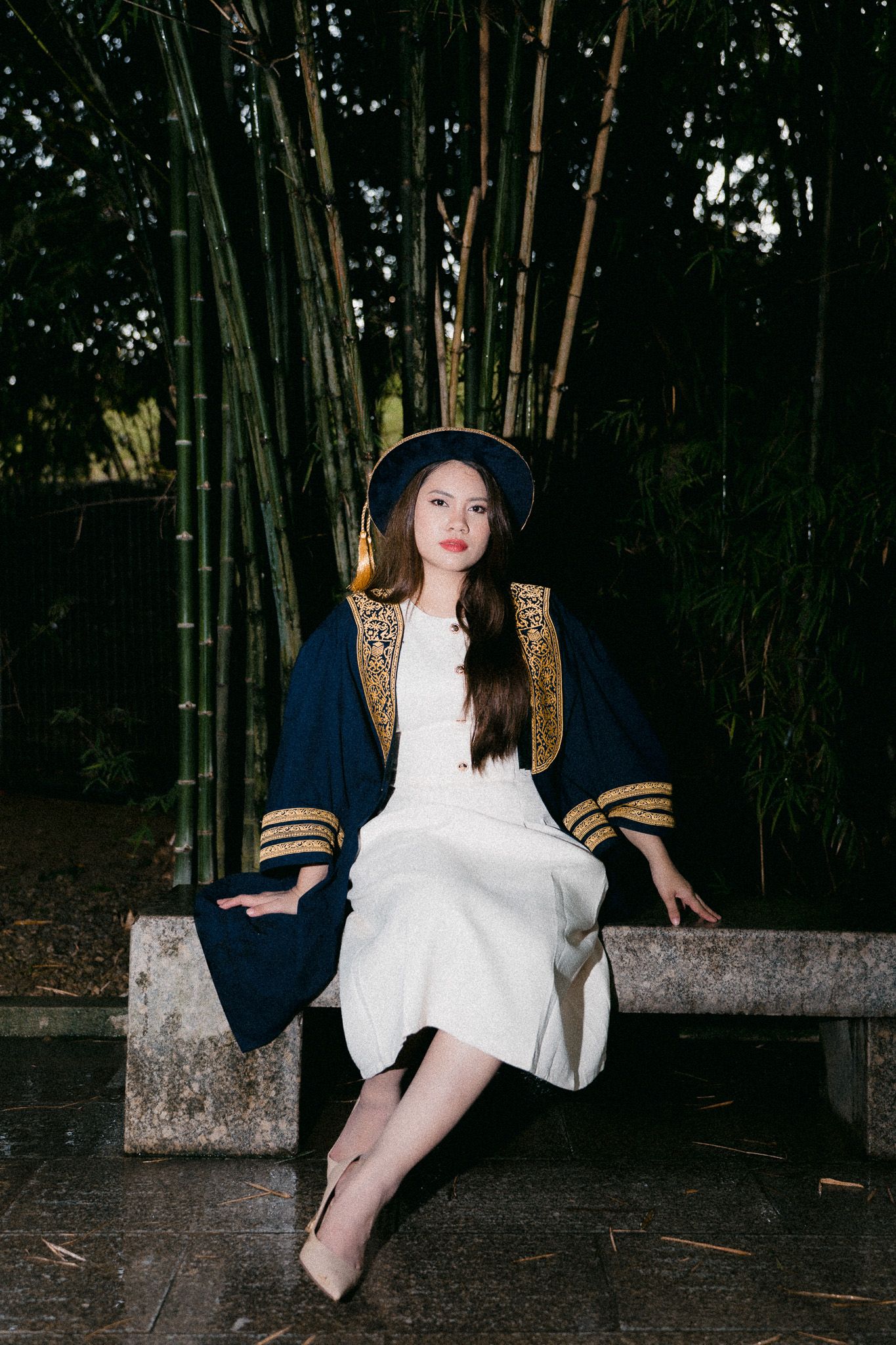 Young Asian woman in navy graduation gown and hat sits on stone bench among bamboo trees.