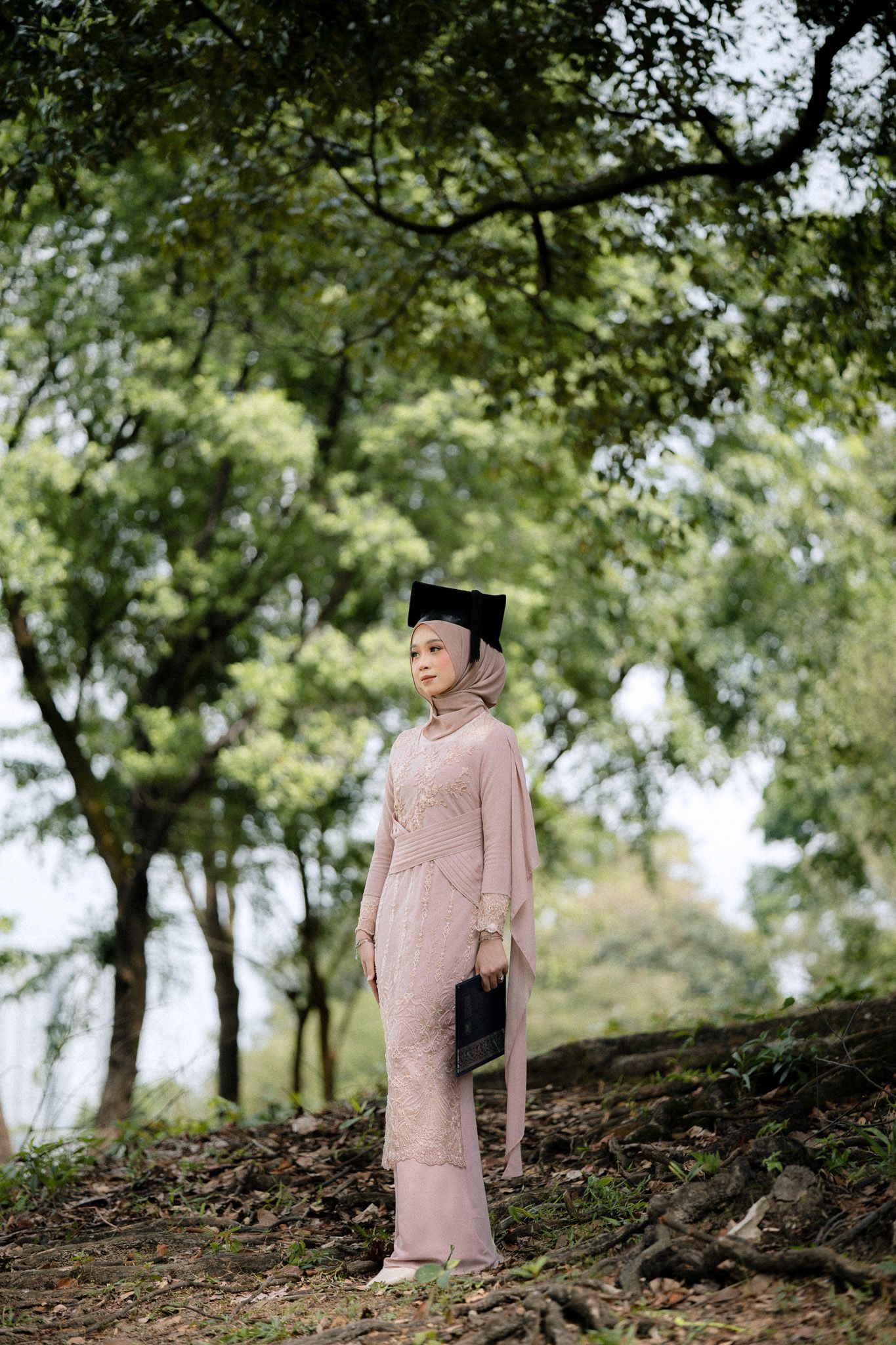 A Muslim woman in a pink kebaya and hijab wearing a graduation cap stands proudly in a lush green forest setting.