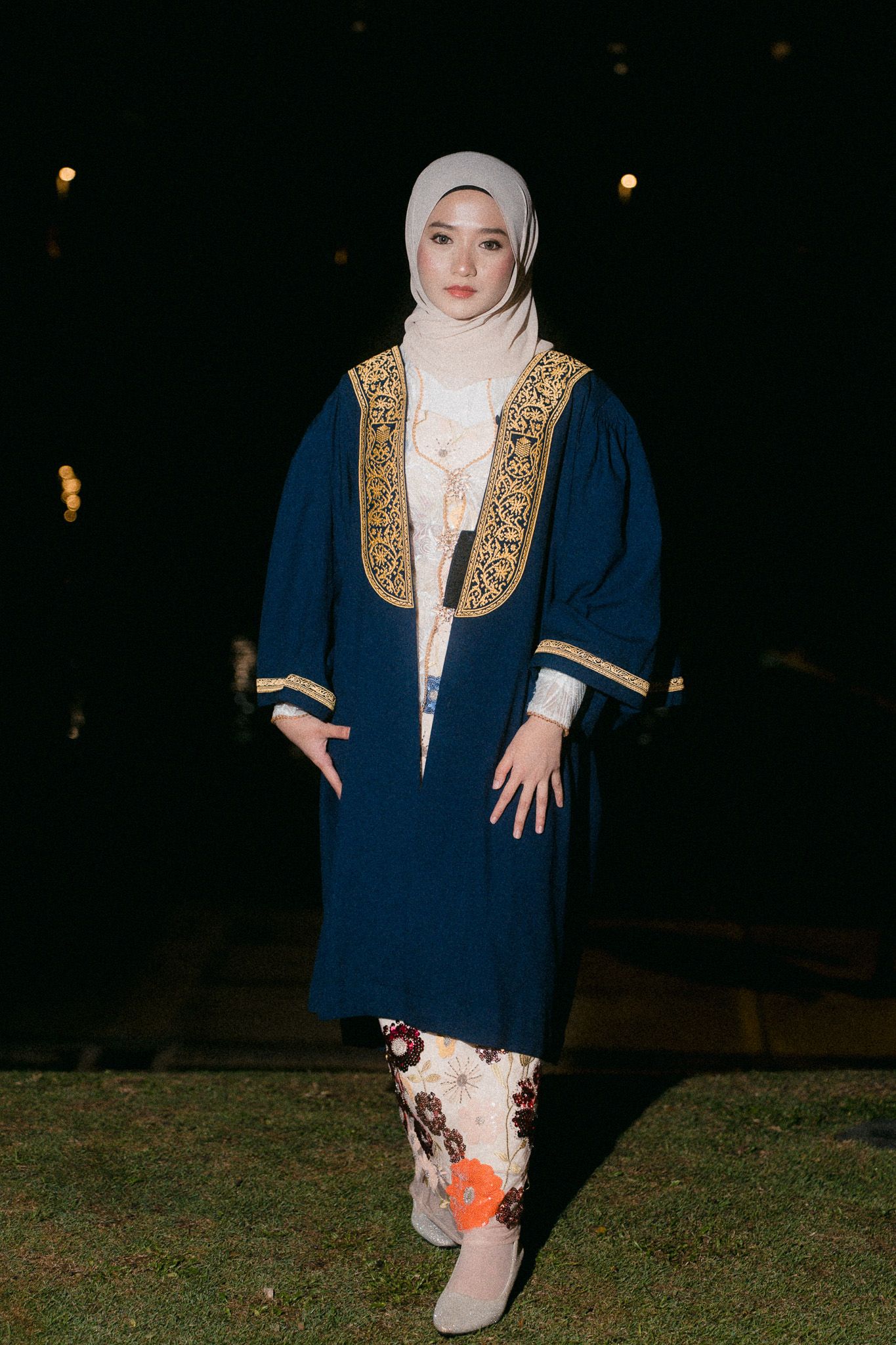 Muslim woman in navy abaya with gold embroidery and floral skirt posing outdoors at night.