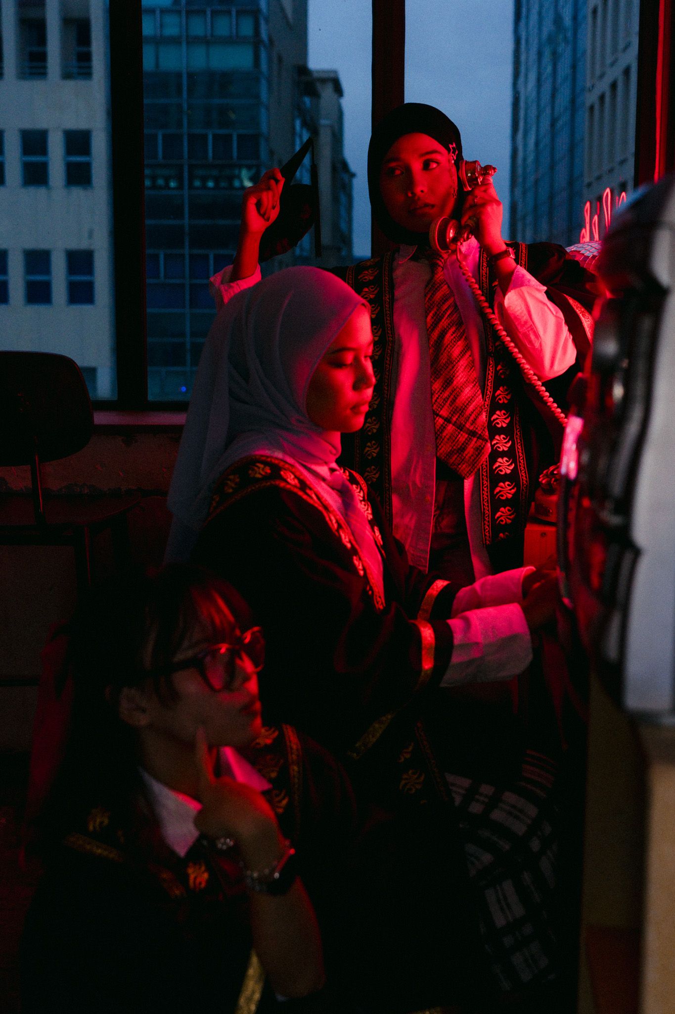 Three women in traditional attire gathered near a window with city views, illuminated by dramatic red lighting.