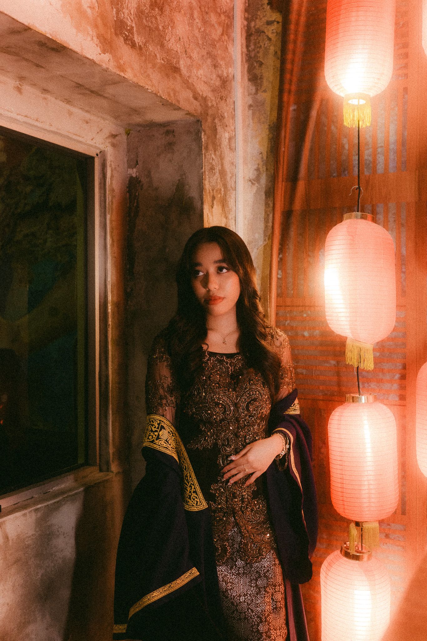 A young woman in an ornate dark embroidered dress poses beside glowing red Chinese lanterns against a textured wall.